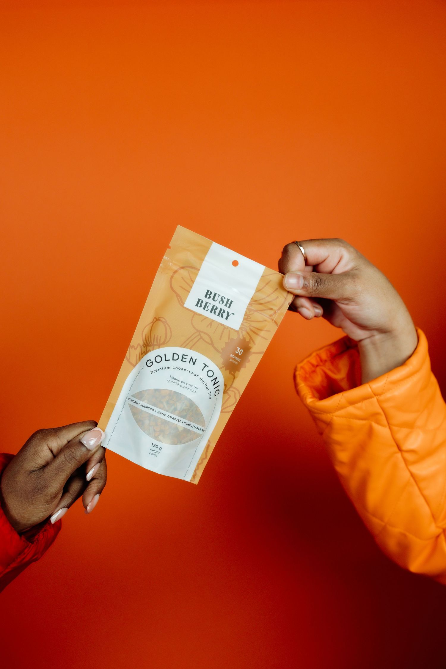 Two hands exchanging a golden product package against a vibrant orange background.