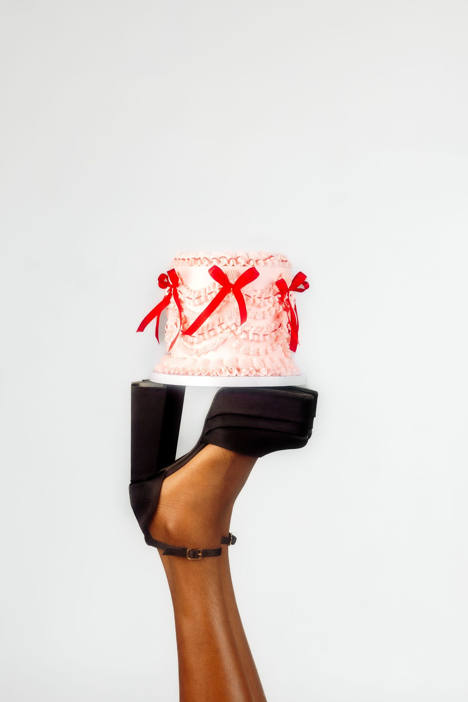 Pink cake decorated with red ribbons held up by a black platform shoe against white background.