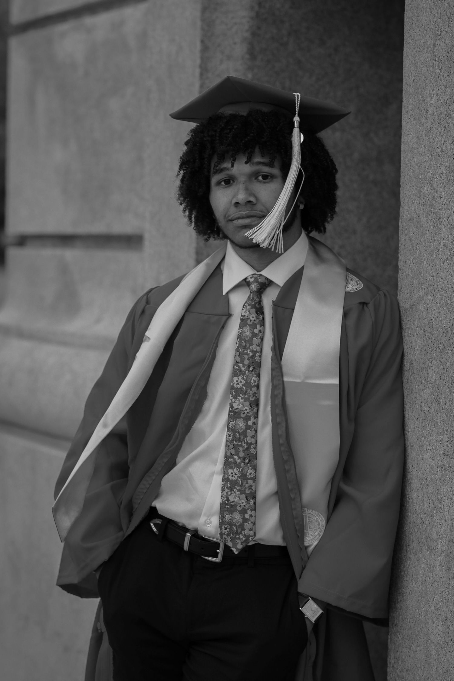Monochrome portrait of graduate in academic regalia against stone wall.