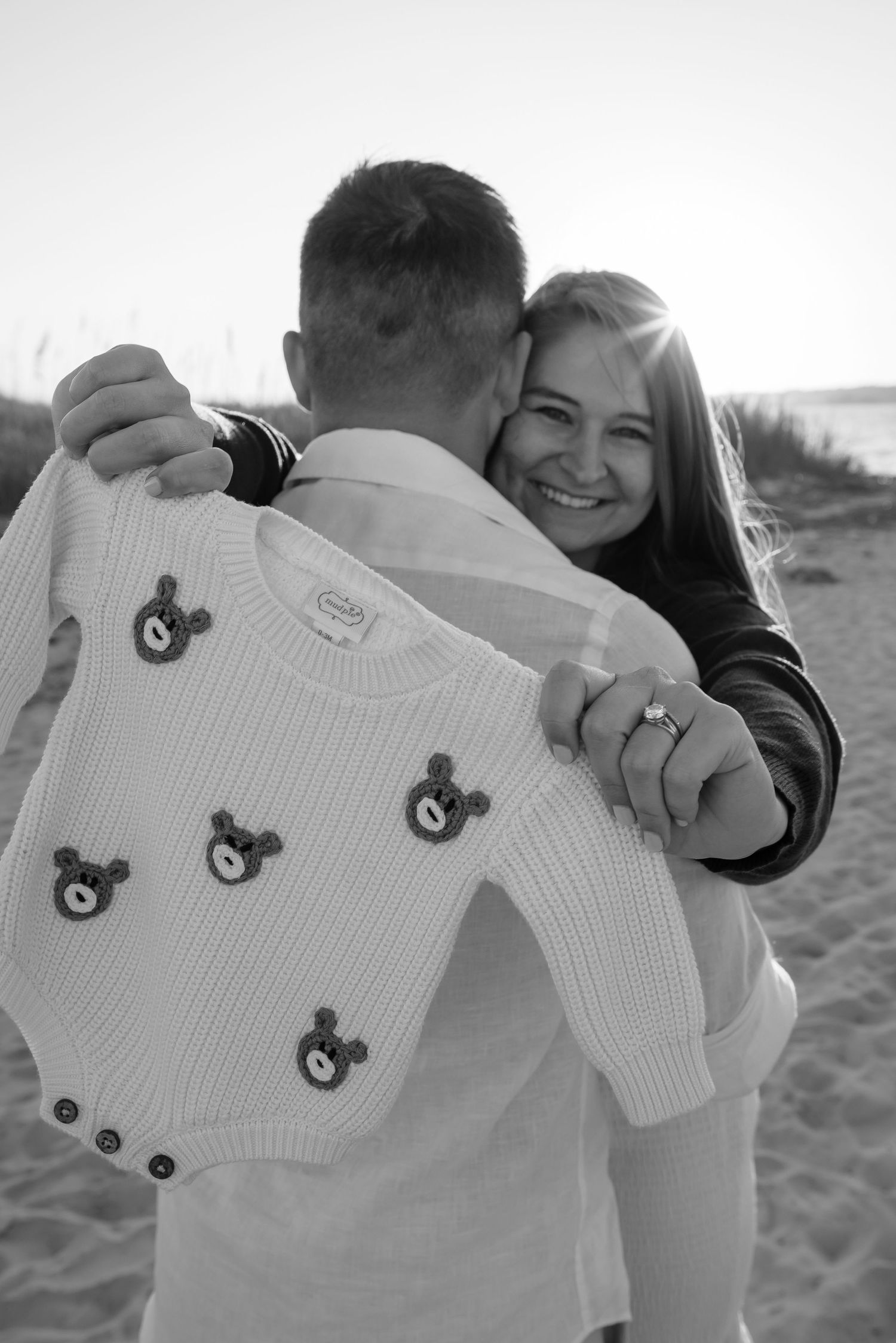 Couple sharing a happy moment while holding a bear-printed baby onesie on the beach.