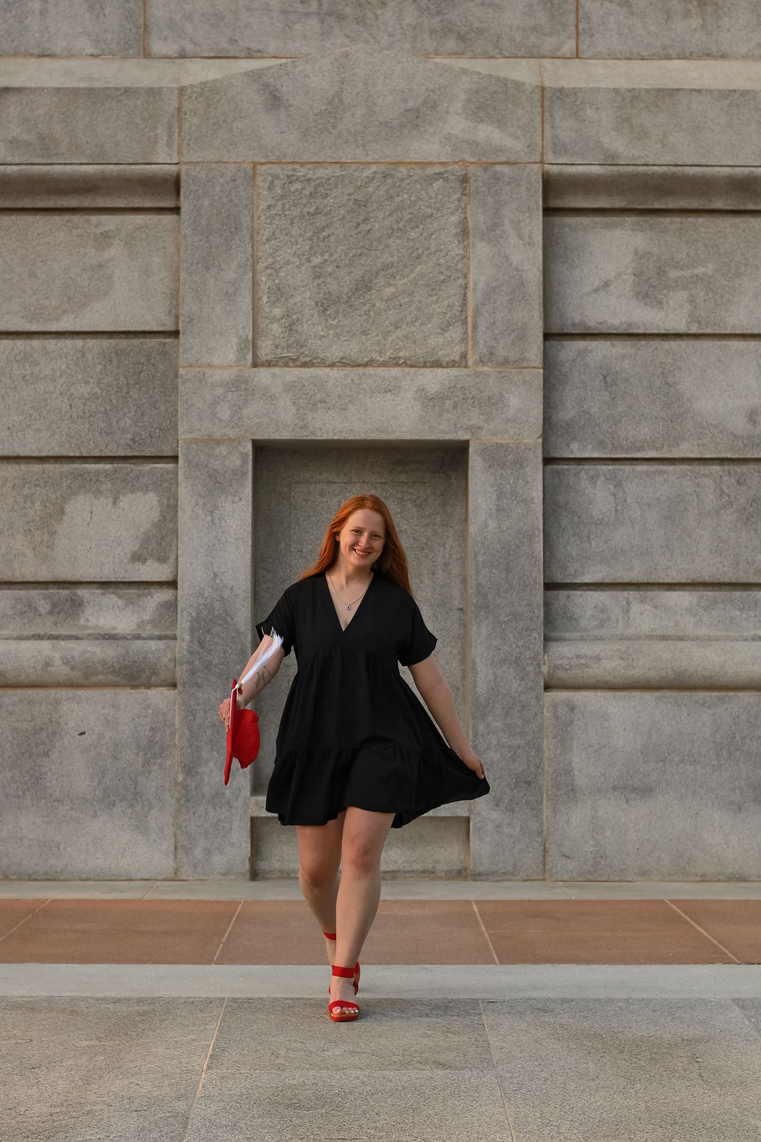 Graduate in black dress and red heels walking confidently against stone architecture.