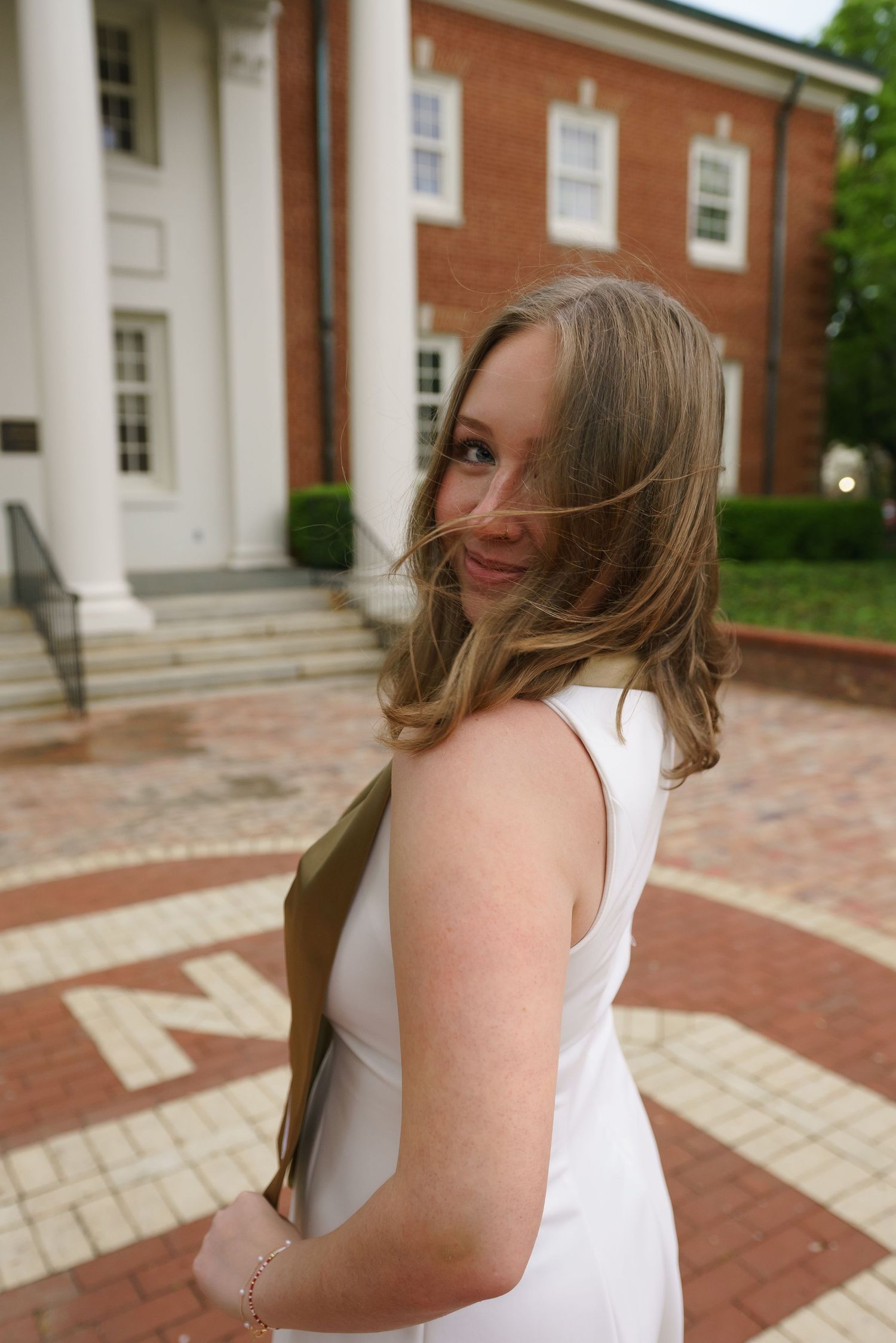 Young woman in white dress smiling over shoulder near brick building with white columns on a windy day.