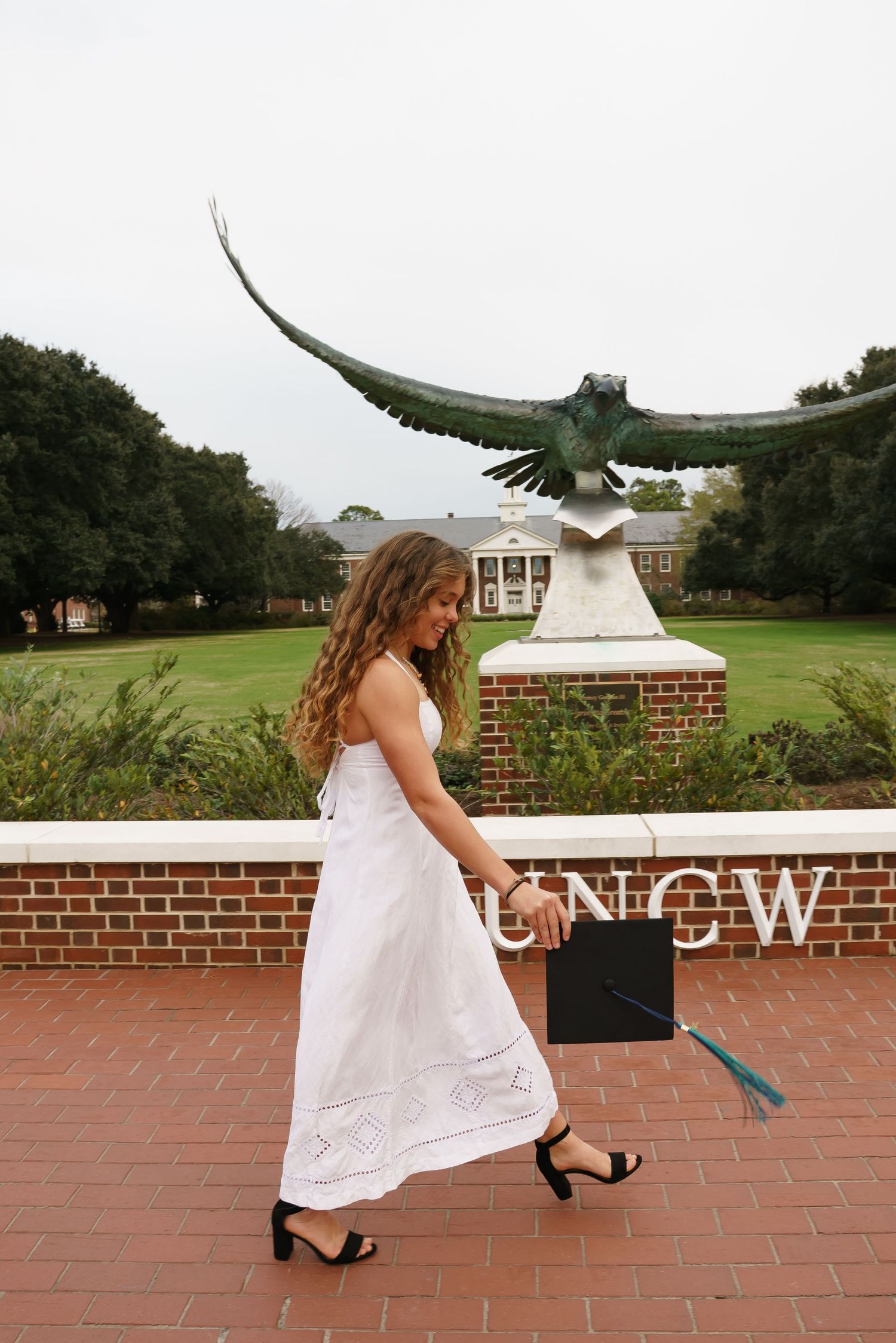 A graduate in white dress pretends to run from a hawk statue at UNCW campus.