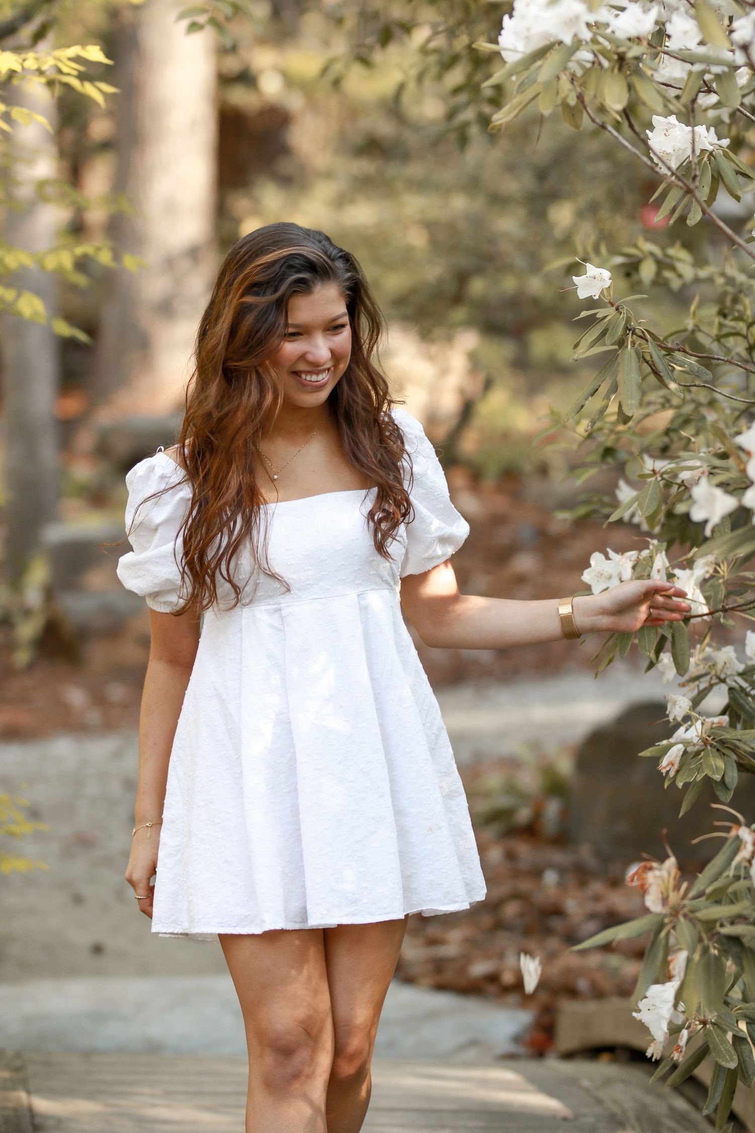 Person in white summer dress walks along garden path near blooming flowers.