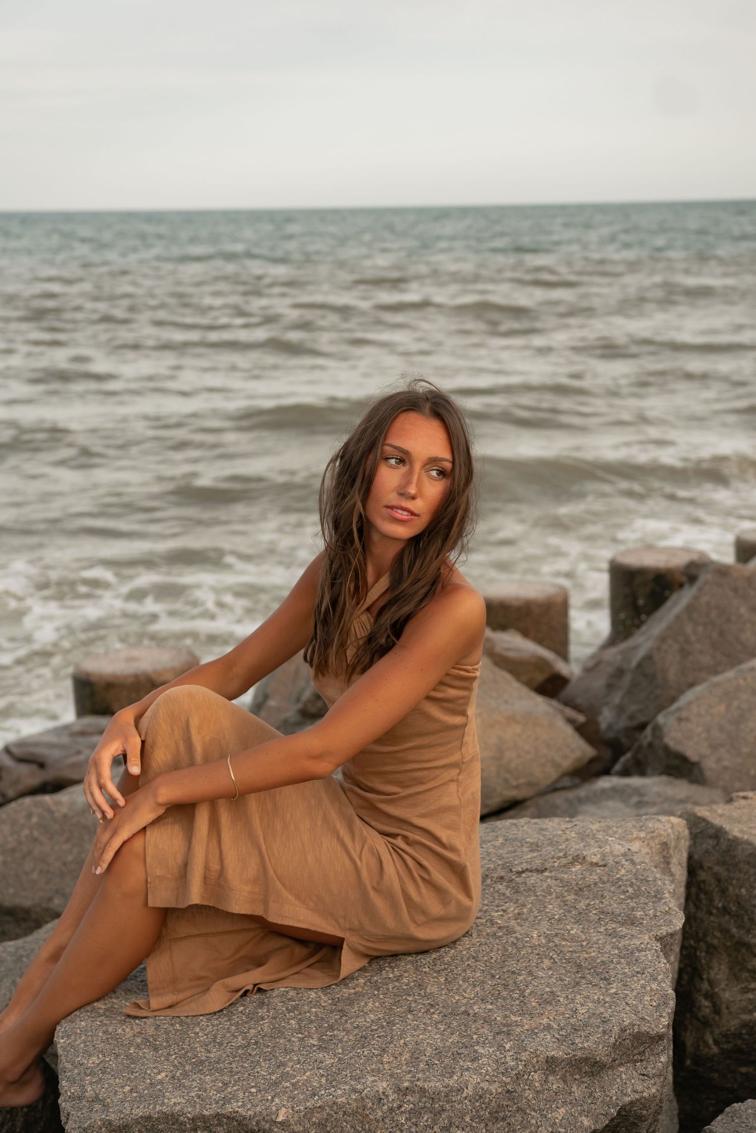 Woman in tan dress poses on rocky beach jetty during golden hour photoshoot.