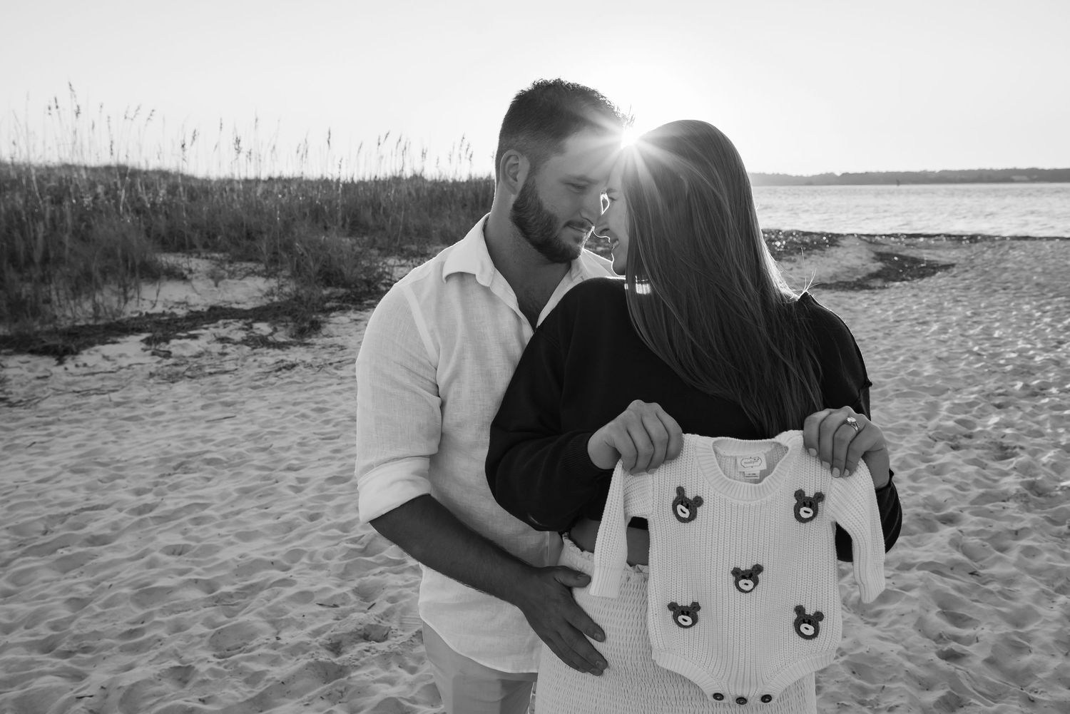 Black and white beach photo of a couple holding a teddy bear printed onesie.