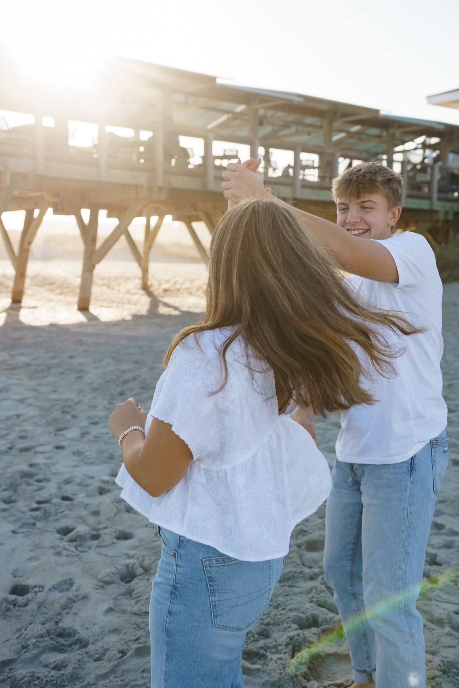 Couple in white shirts and jeans dancing playfully on beach near wooden pier.