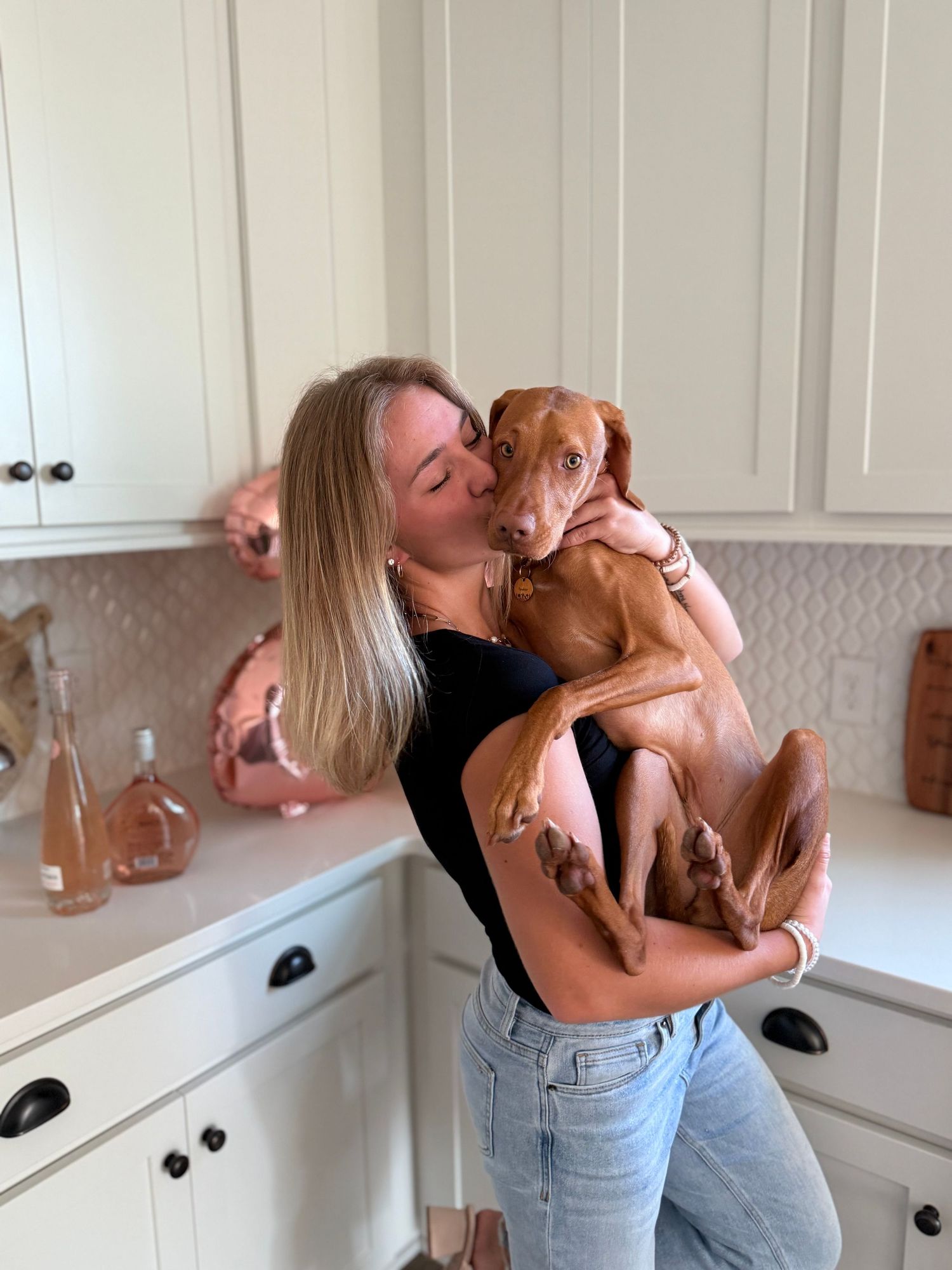 Woman kissing her brown Vizsla puppy in a bright white kitchen with rose gold decor.
