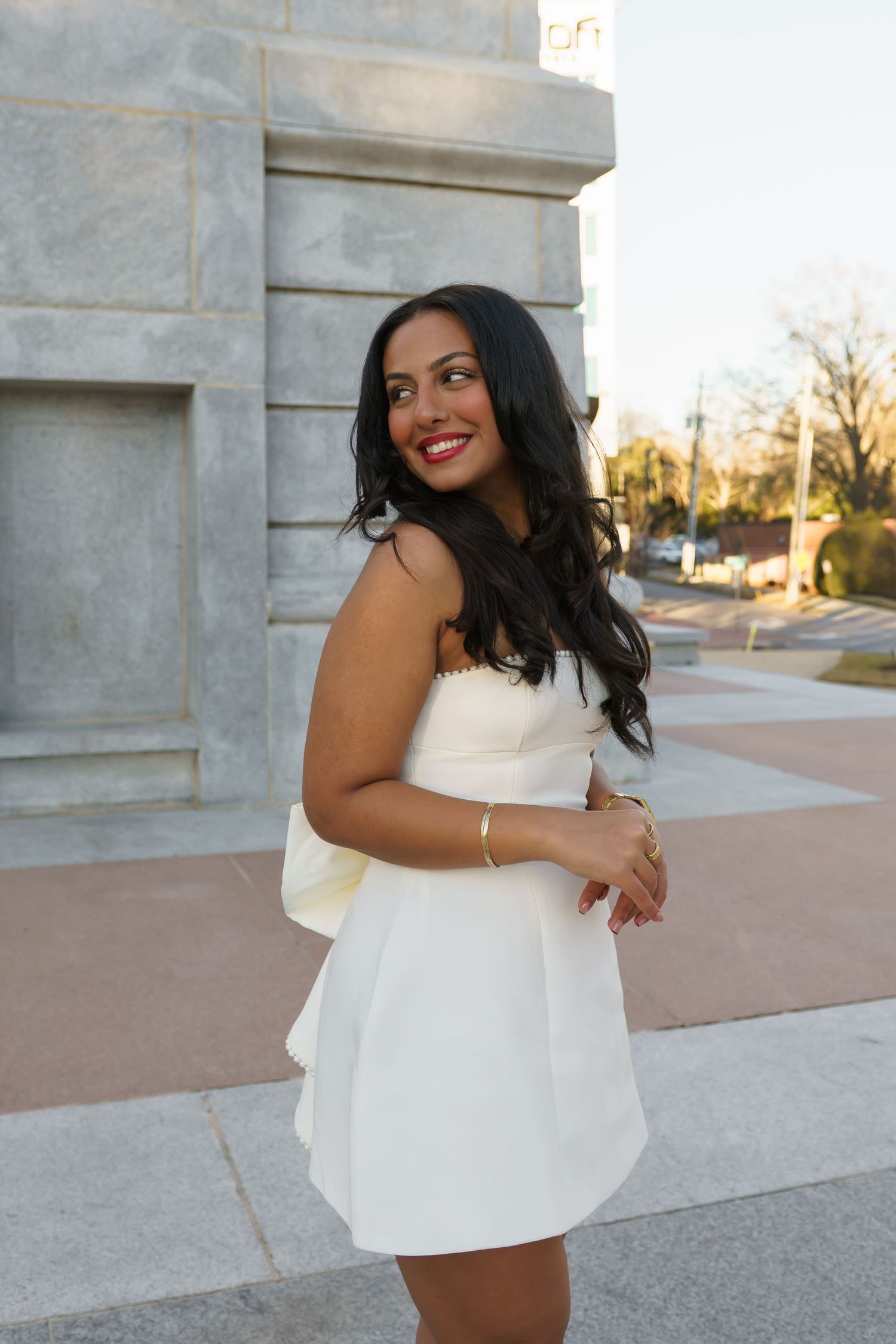 Young woman in white strapless mini dress smiling outdoors near stone columns, carrying a white bag.