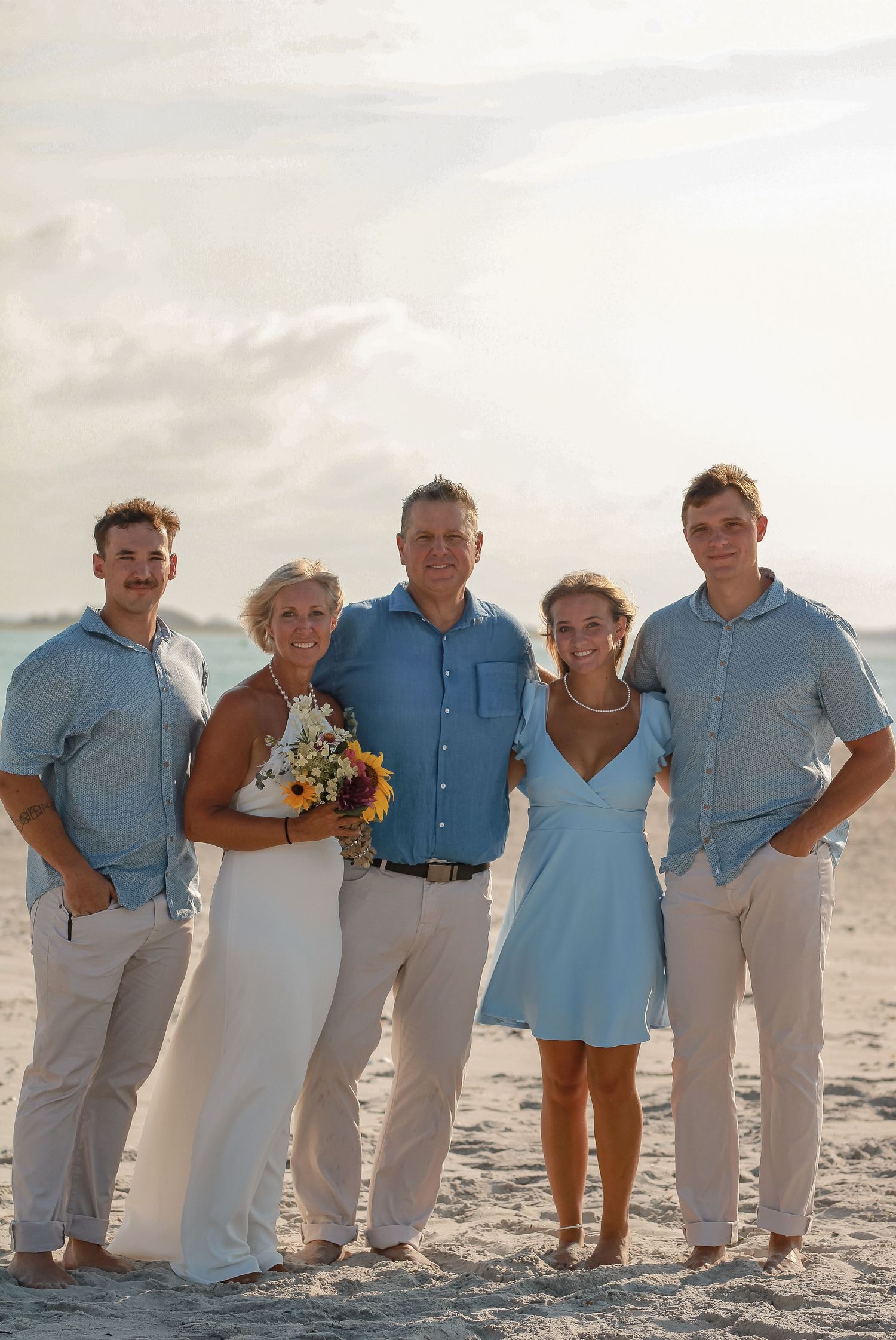 Family group in coordinated blue and white outfits posing on beach at sunset.