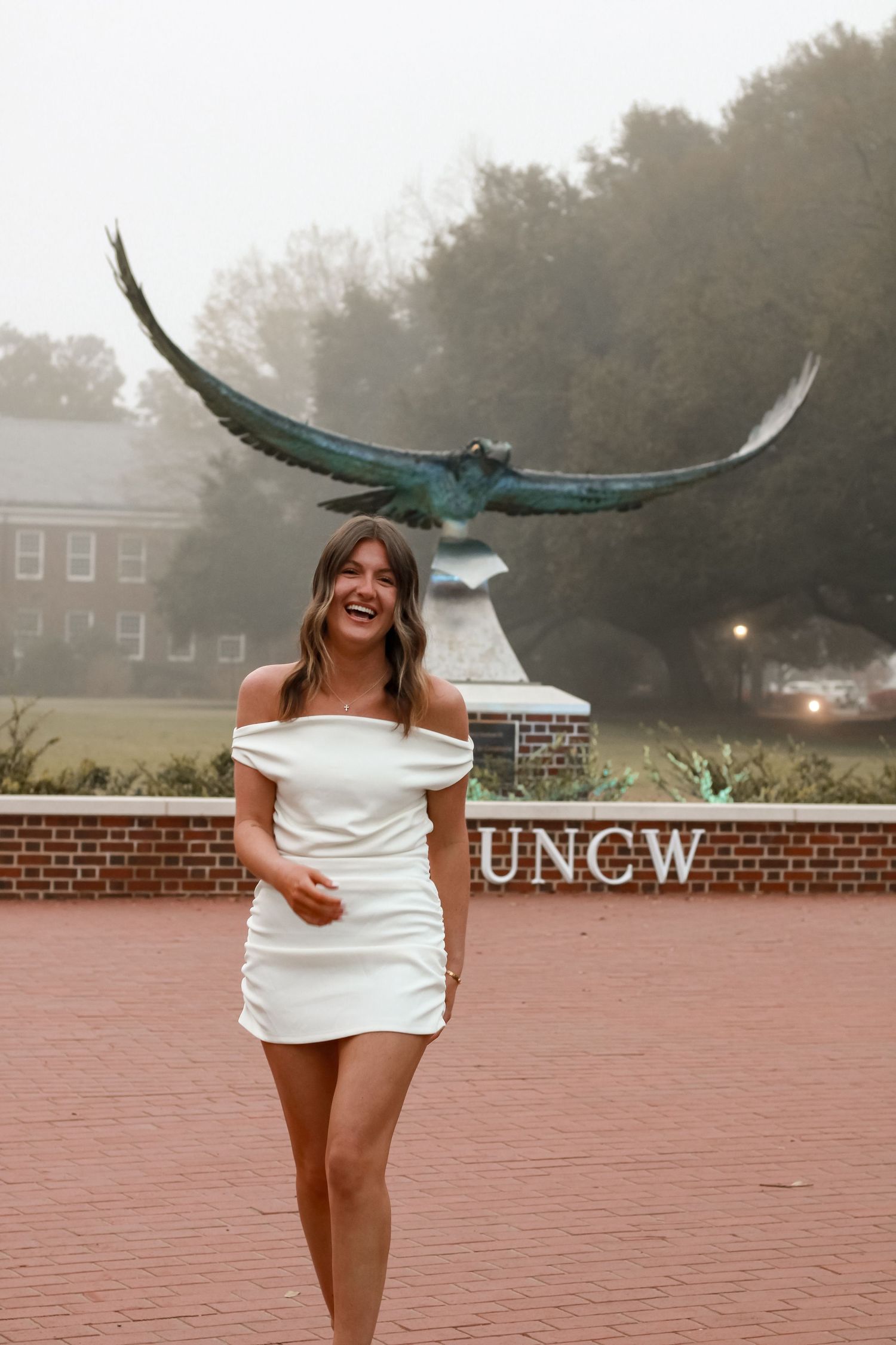 Graduate posing in white dress by UNCW eagle statue on foggy day.