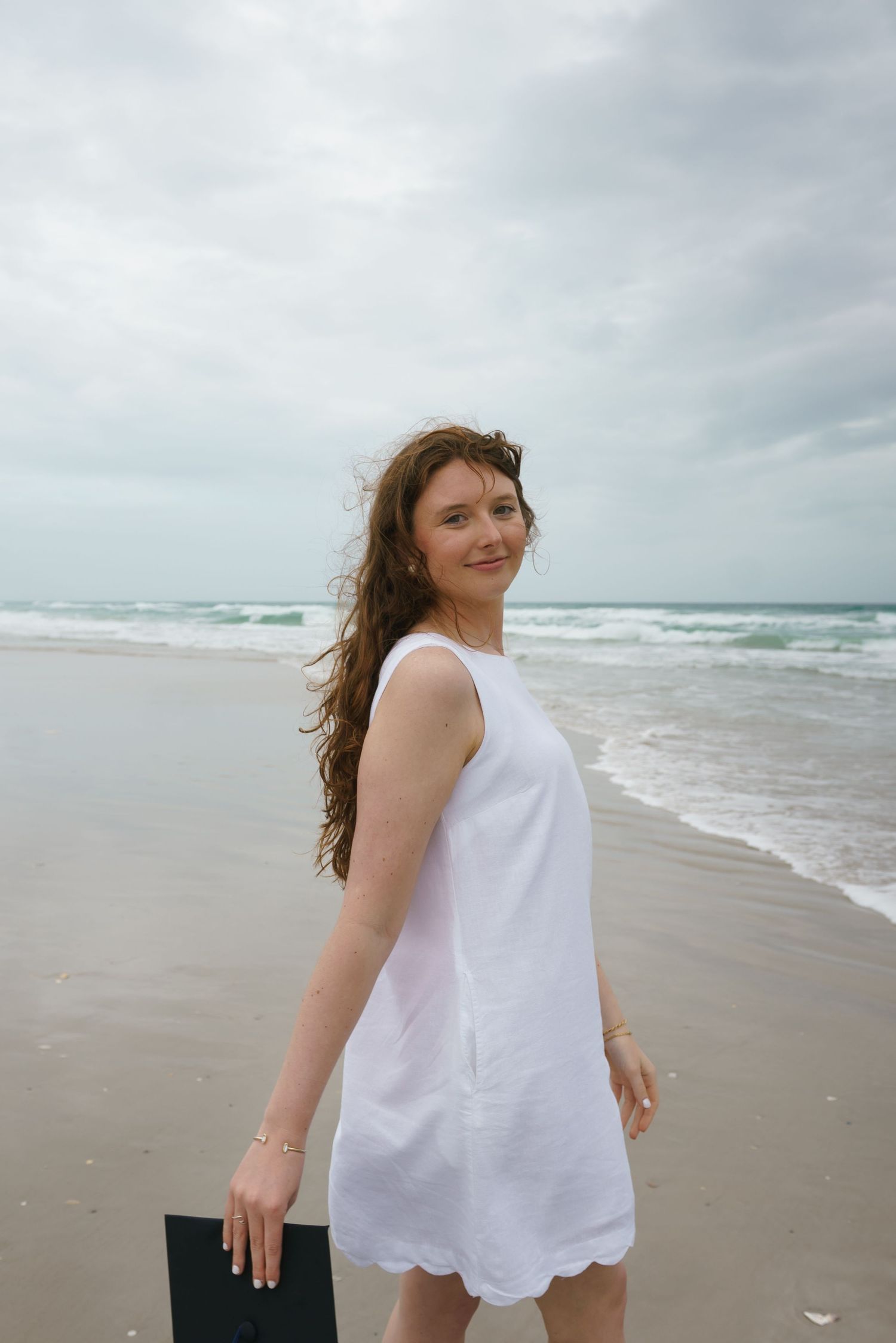 Young woman in white sleeveless dress smiling on a sandy beach with ocean waves behind her.