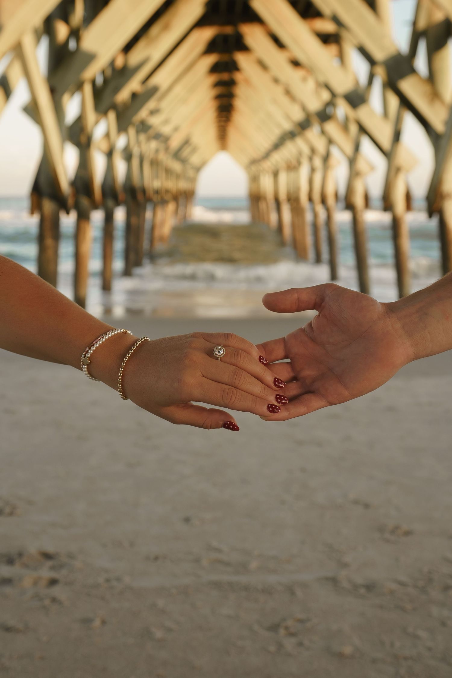 Two hands reaching towards each other under a pier, showing jewelry and engagement rings.
