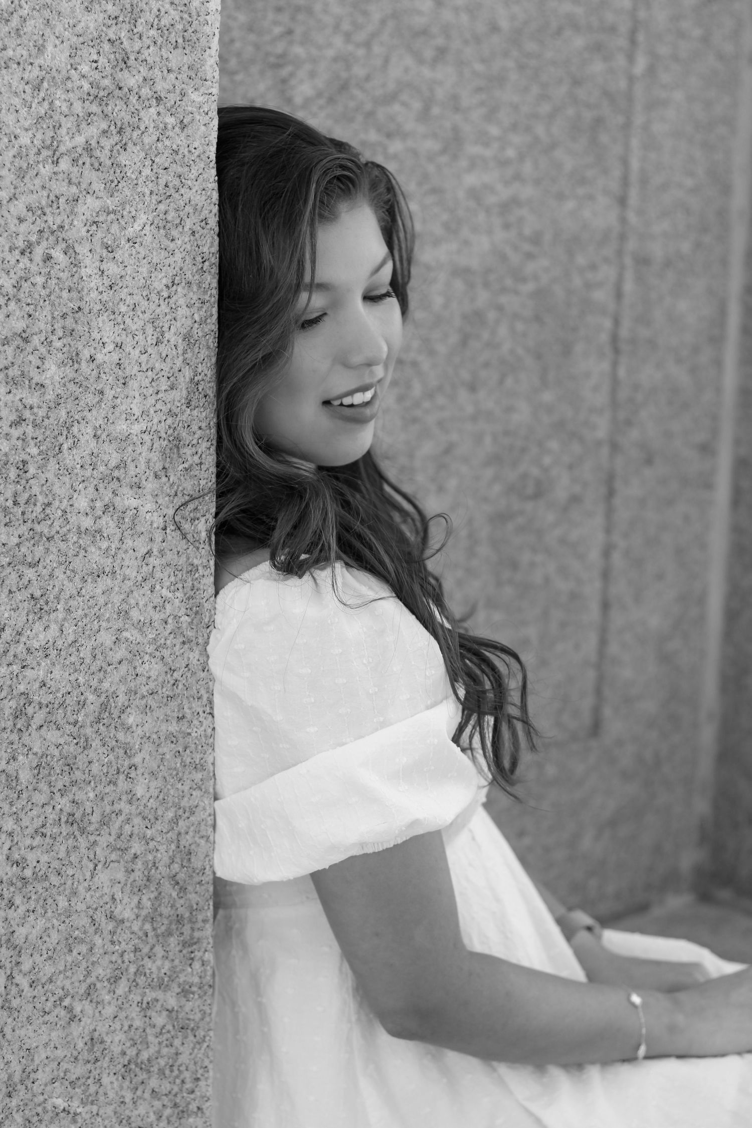 Black and white portrait of person peeking from behind stone column while wearing white dress.