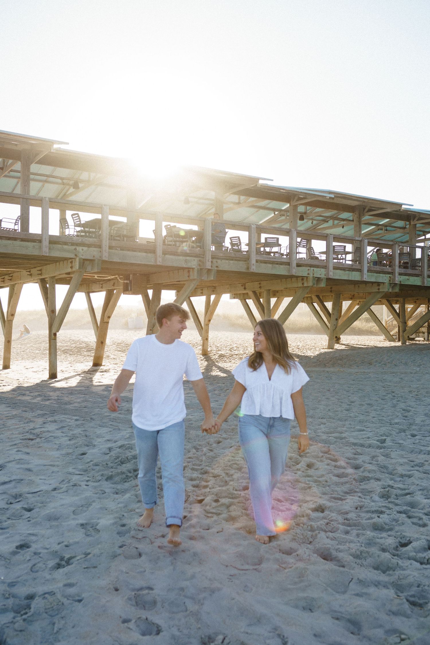 Couple walking barefoot on sandy beach at sunset under wooden pier.