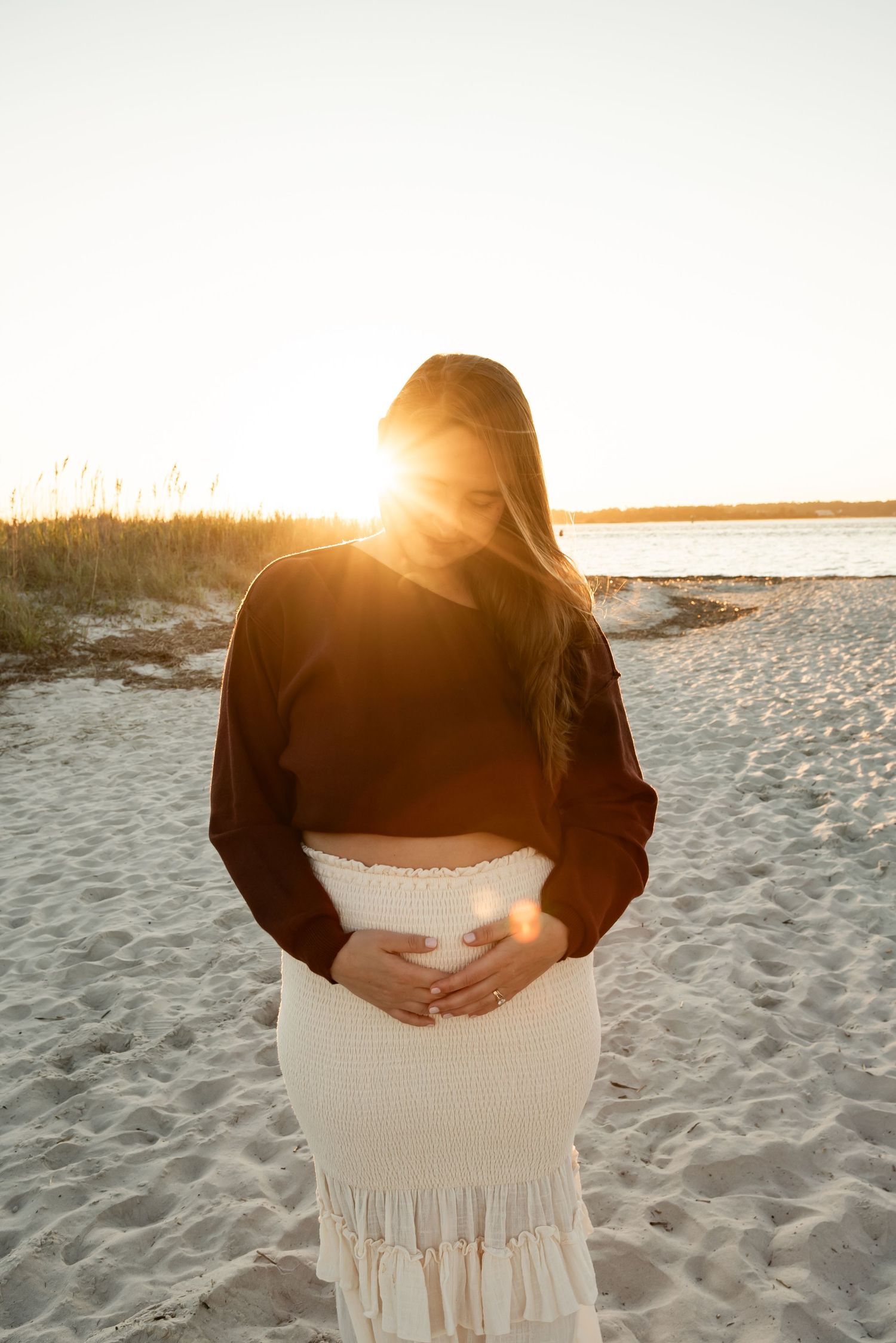 A person in a black sweater stands on a beach at sunset holding their pregnant belly.