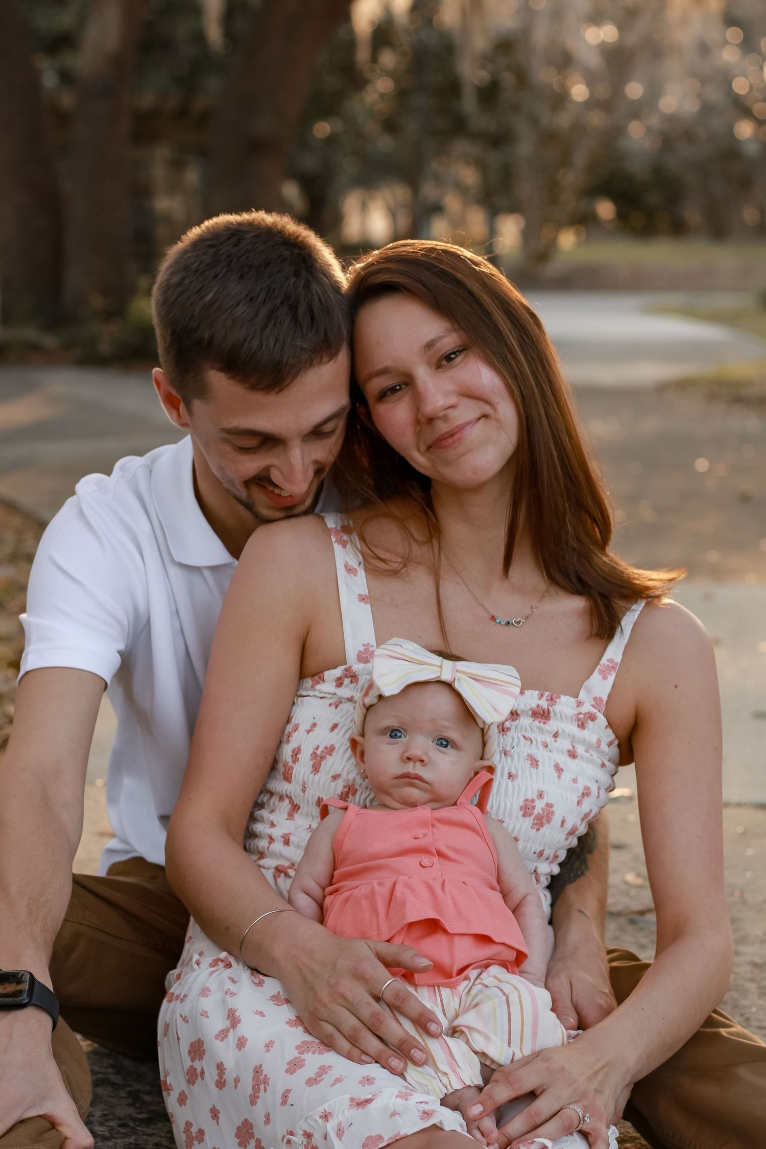 A young family poses together outdoors with their baby in a floral dress and coral outfit during golden hour.