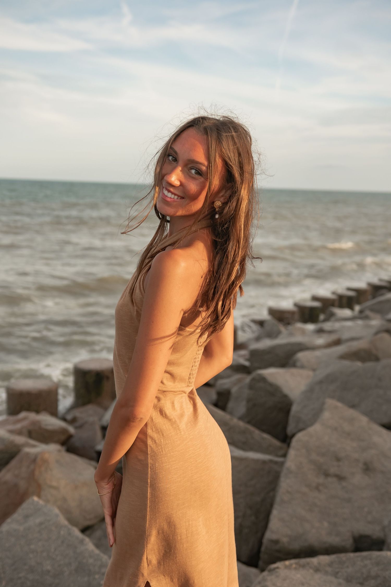 Woman in beige dress smiles while posing on coastal rocks at sunset.
