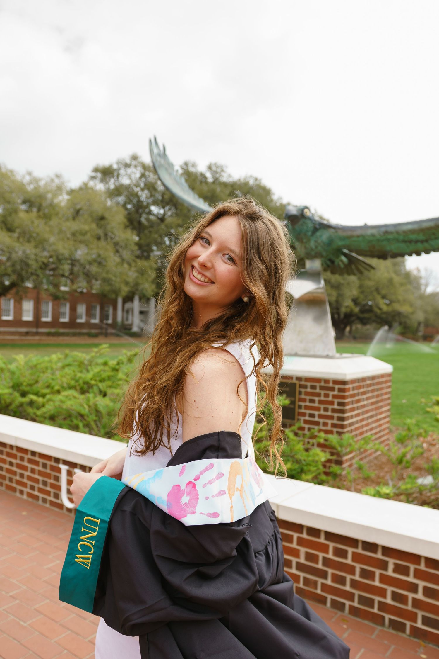 Young woman smiling in UNCW graduation regalia outdoors near a bird sculpture on a college campus.
