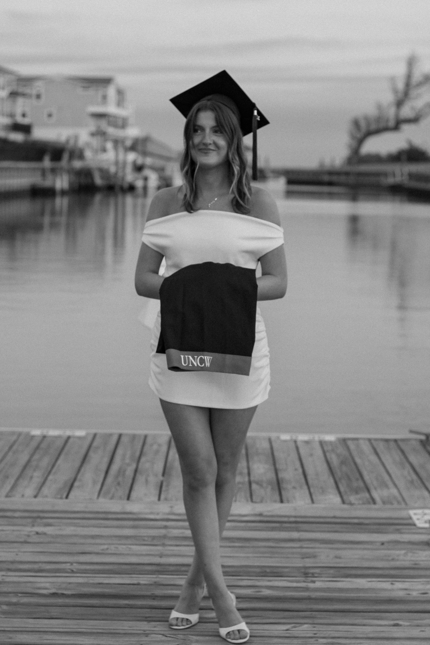 Black and white photo of graduate in cap and gown standing on wooden dock by water.
