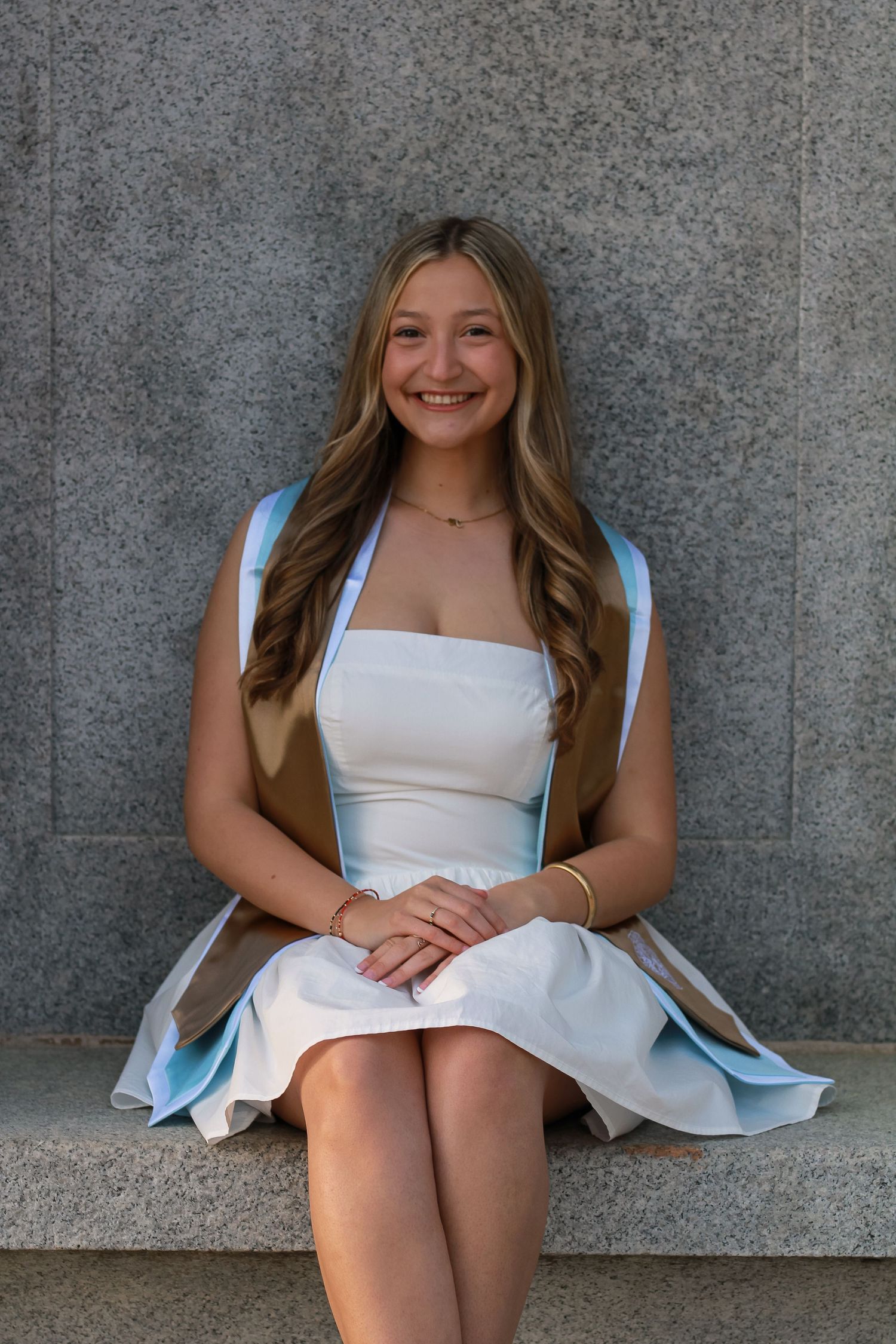 Person in a white dress sitting against a gray wall in a casual pose.
