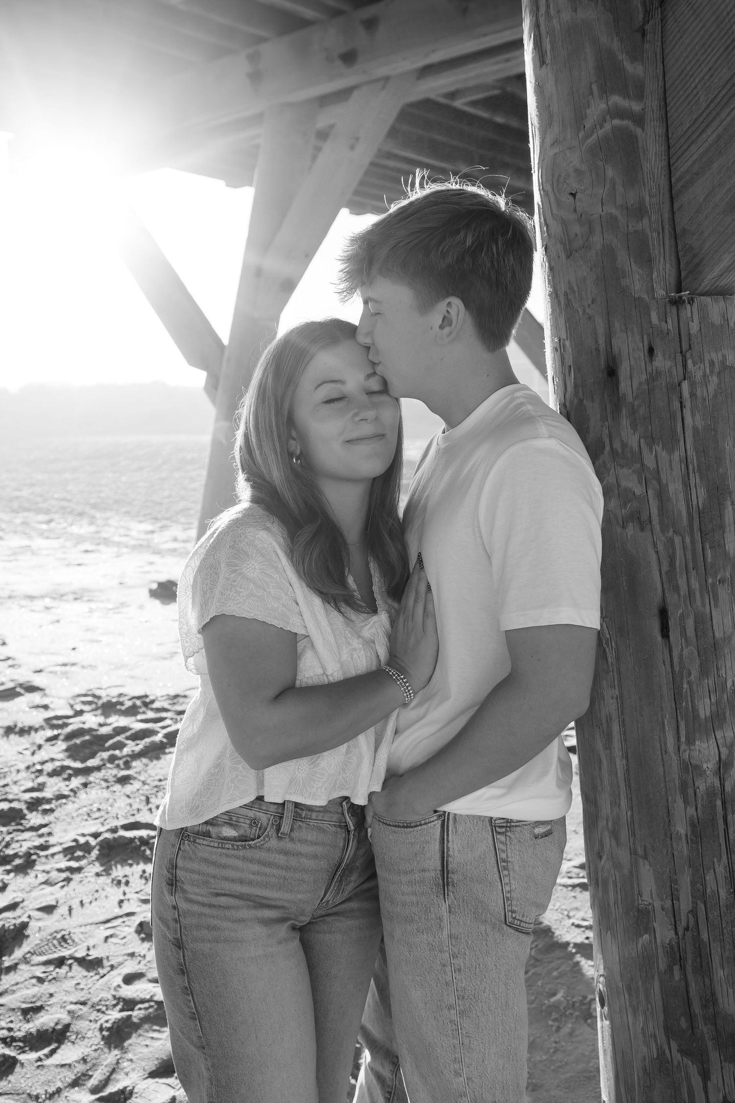 Romantic couple embracing against wooden pier posts in dramatic beach lighting.