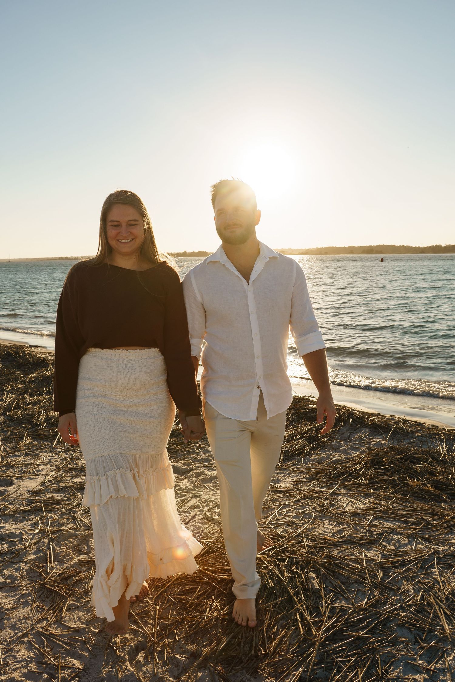 Couple in white outfits walking along beach at sunset.
