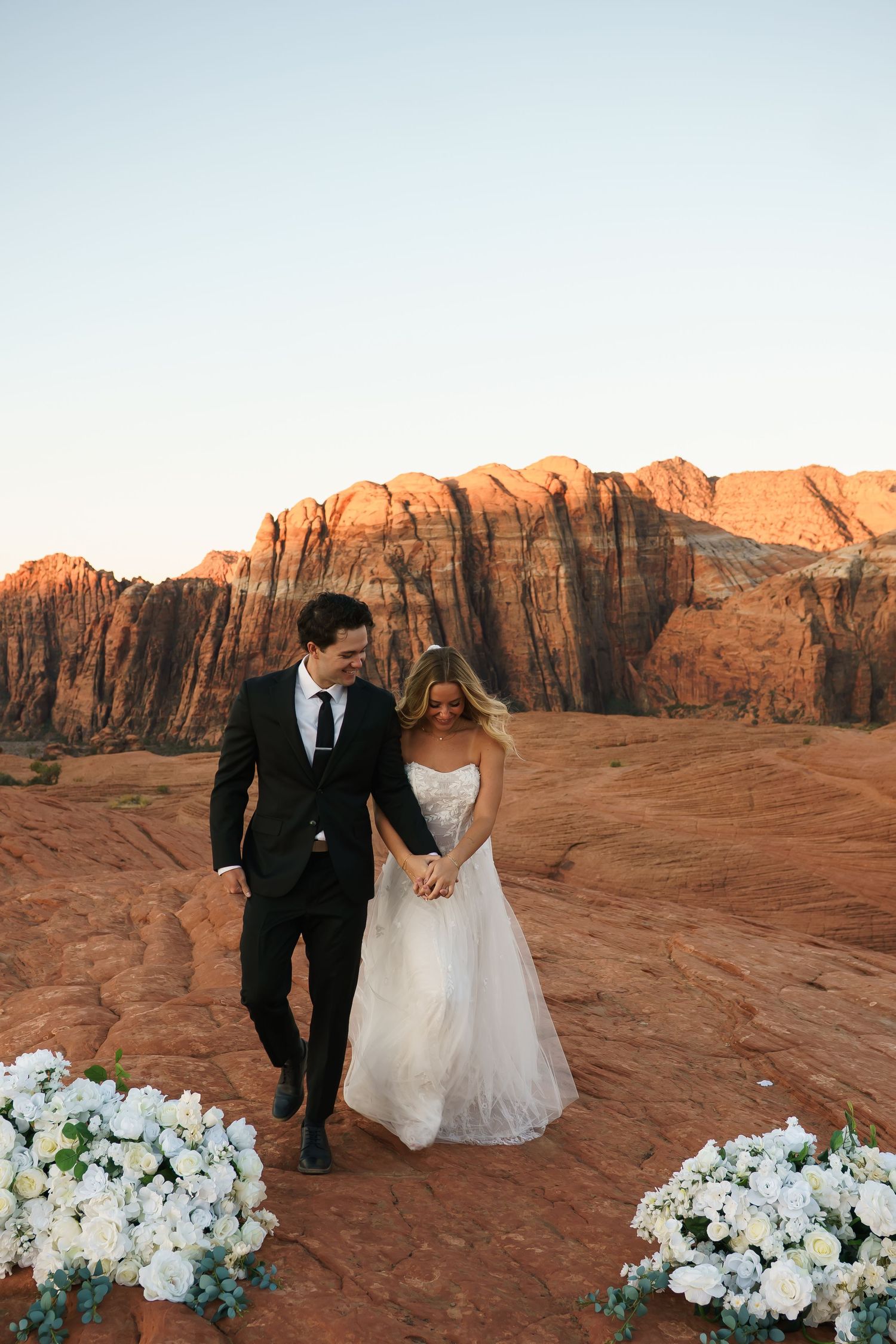 A couple in wedding attire walks hand in hand across red rock formations at sunset with white floral arrangements.