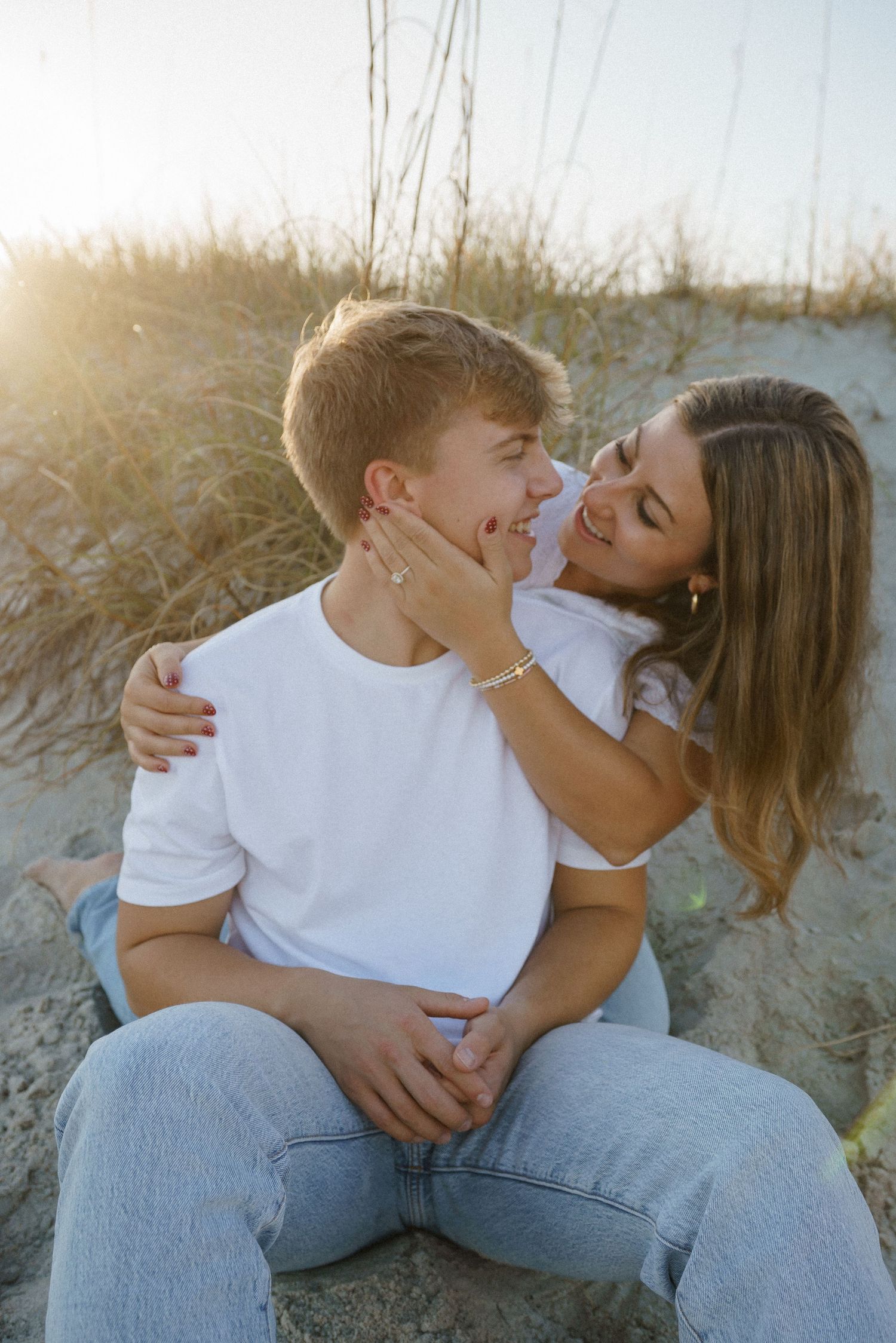 Playful couple laughing and cuddling on sandy beach dunes during golden hour.