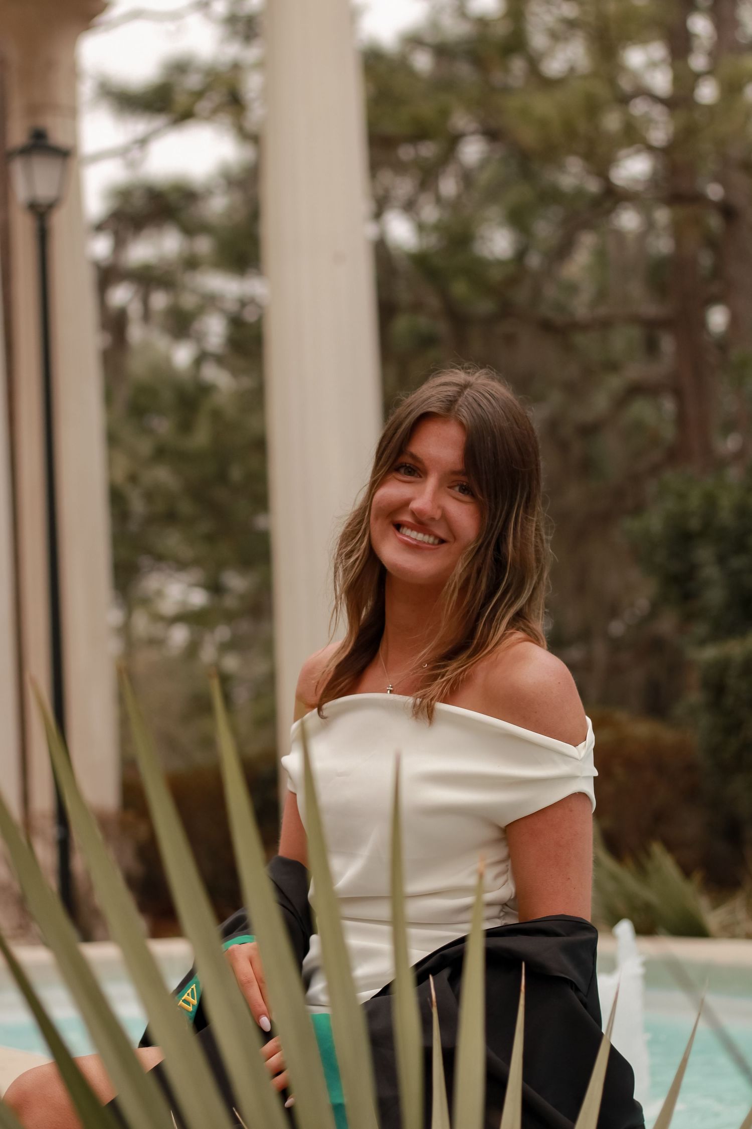 Graduate in off-shoulder white dress poses by classical columns with tropical plants.