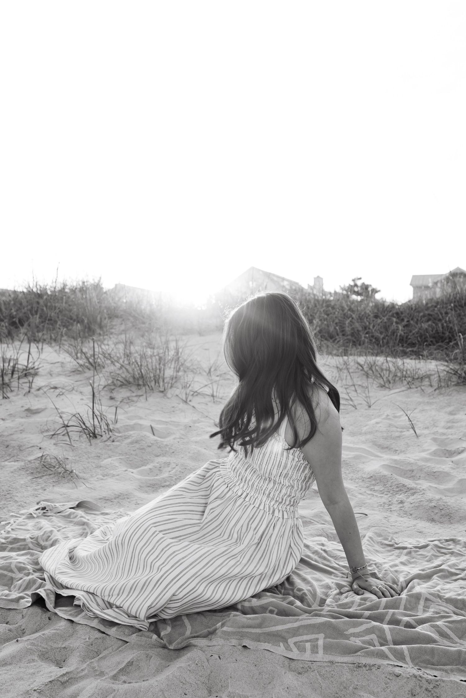 Black and white photo of a woman in a striped dress sitting on a sandy beach blanket, facing away toward bright sunlight.