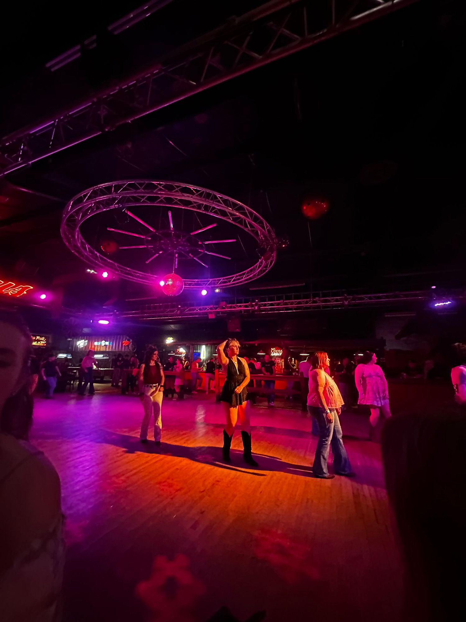 People dancing on a wooden floor in a dimly lit country western bar with purple stage lighting and circular truss fixture.