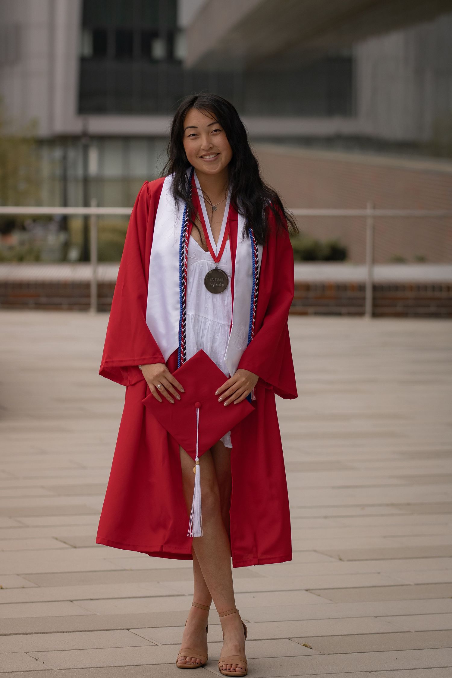 Graduate in red academic regalia and white dress poses on outdoor plaza.