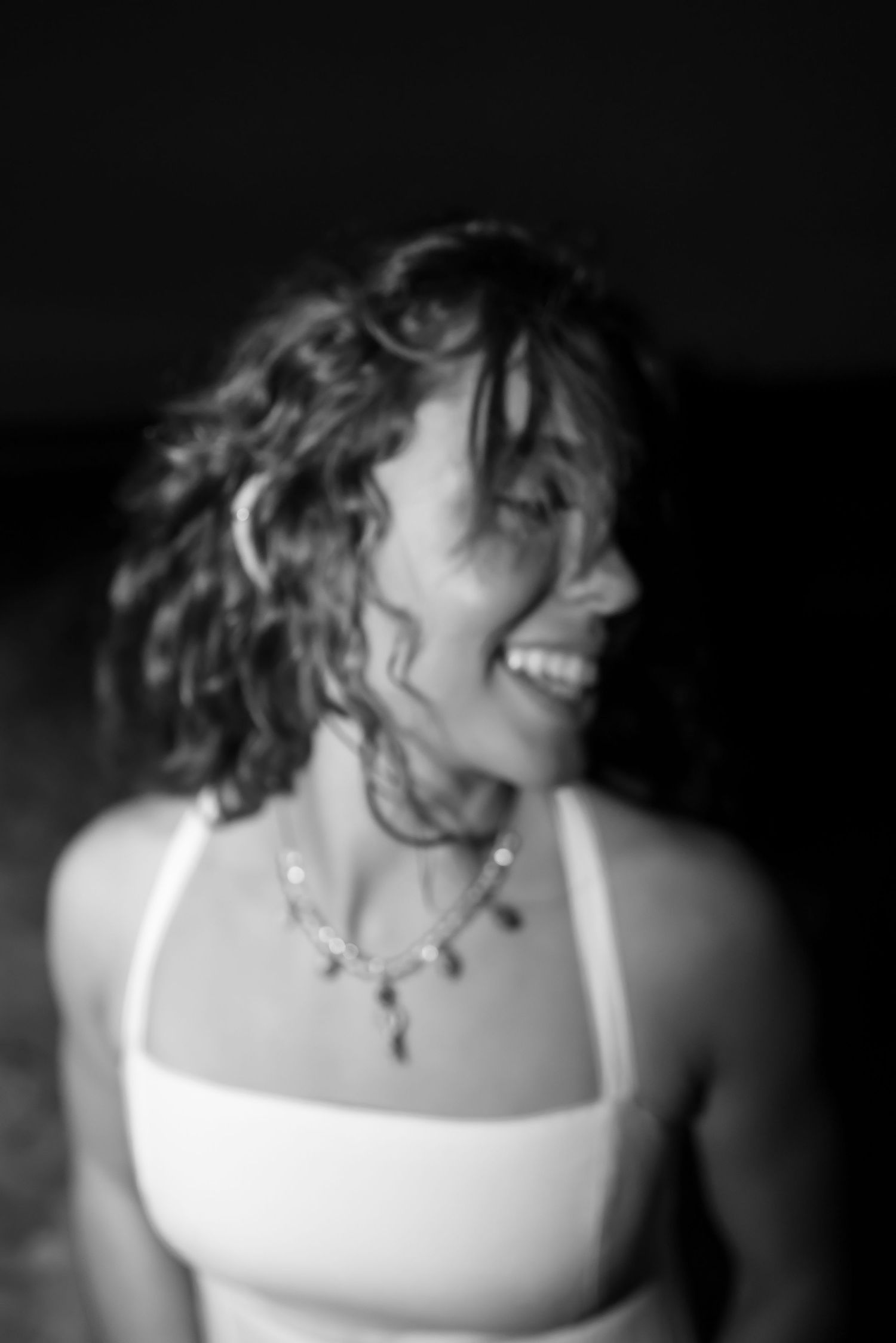 Black and white portrait of a smiling woman with curly hair wearing a white top and delicate necklace.