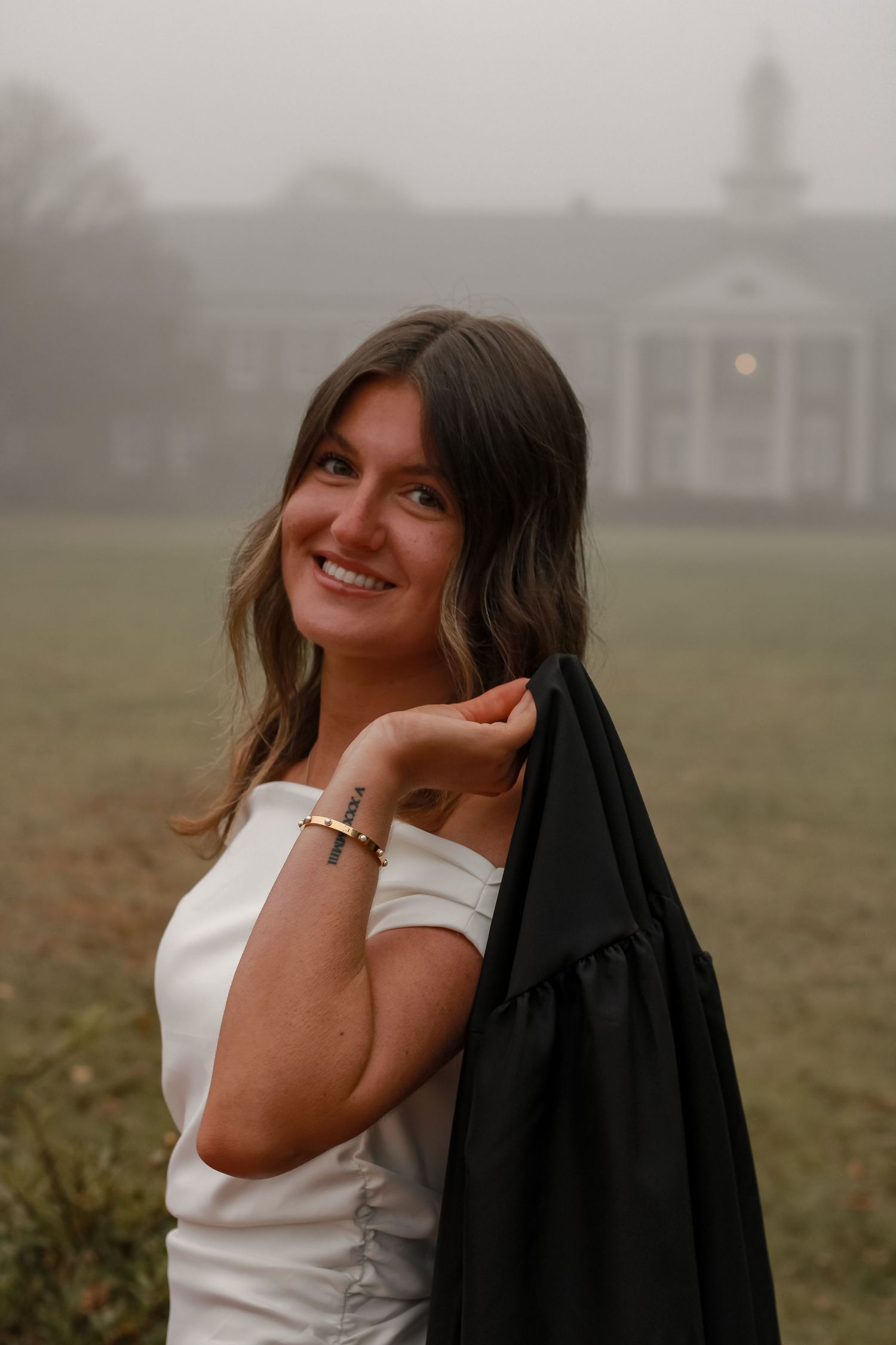 A graduate poses in a white dress and black jacket on a misty campus lawn with a building in the background.