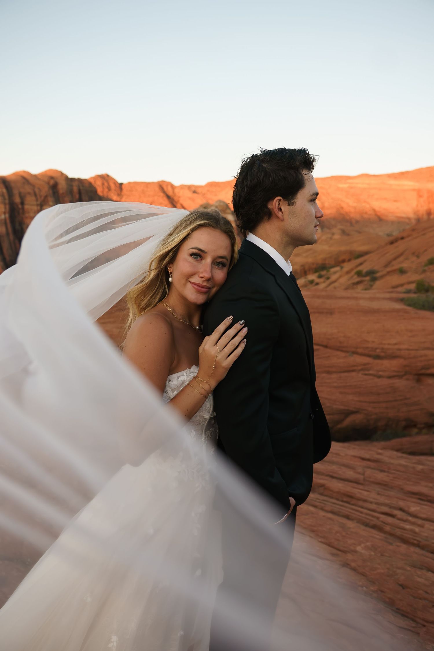 Bride's veil flows dramatically in desert wind as couple embraces on red rocks.