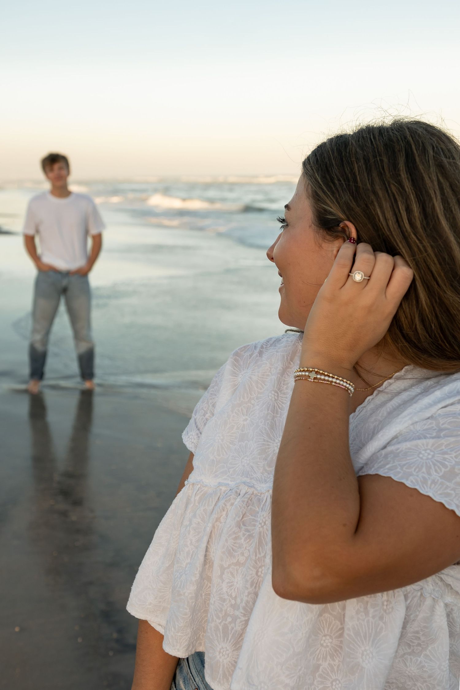 A woman touches her hair while looking at an engagement ring with her partner in the background.