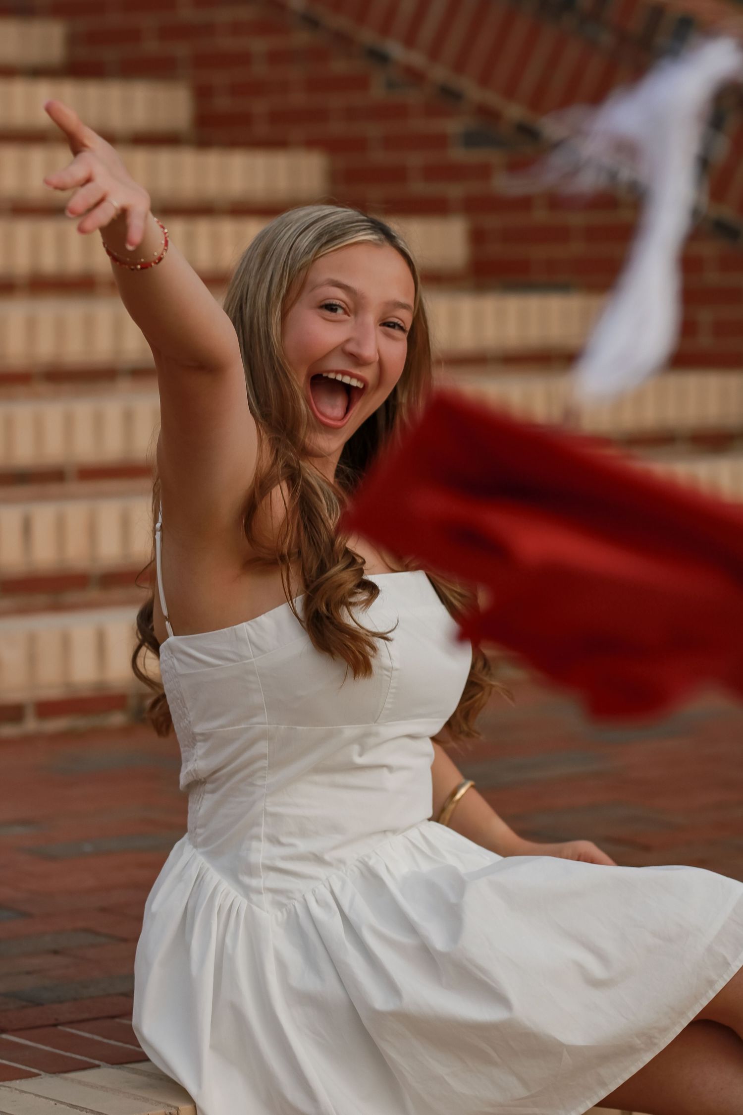 Joyful graduate in white dress tossing red graduation cap on brick steps.