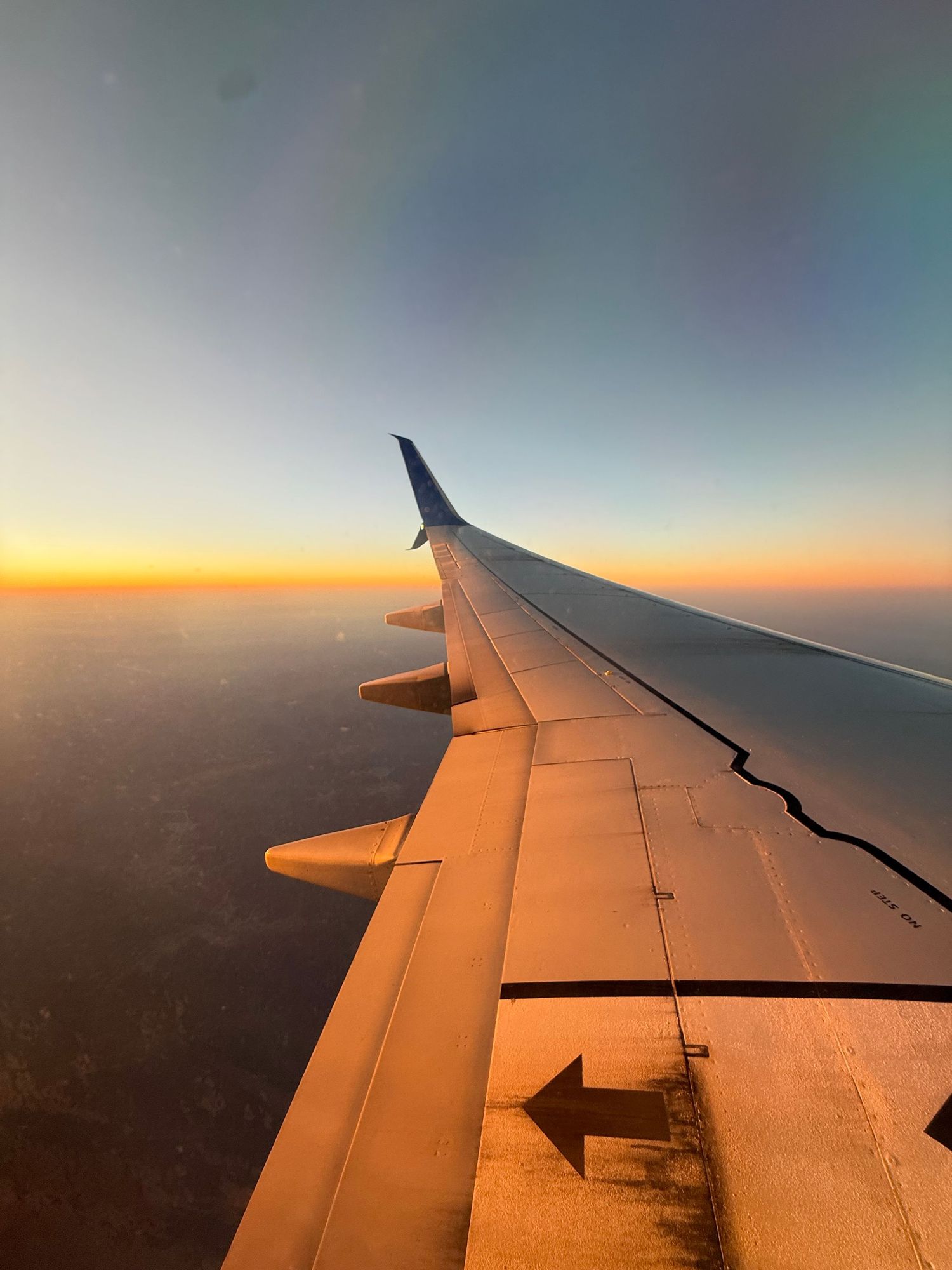 Airplane wing view at golden hour sunset with warm orange sky and vast landscape below.