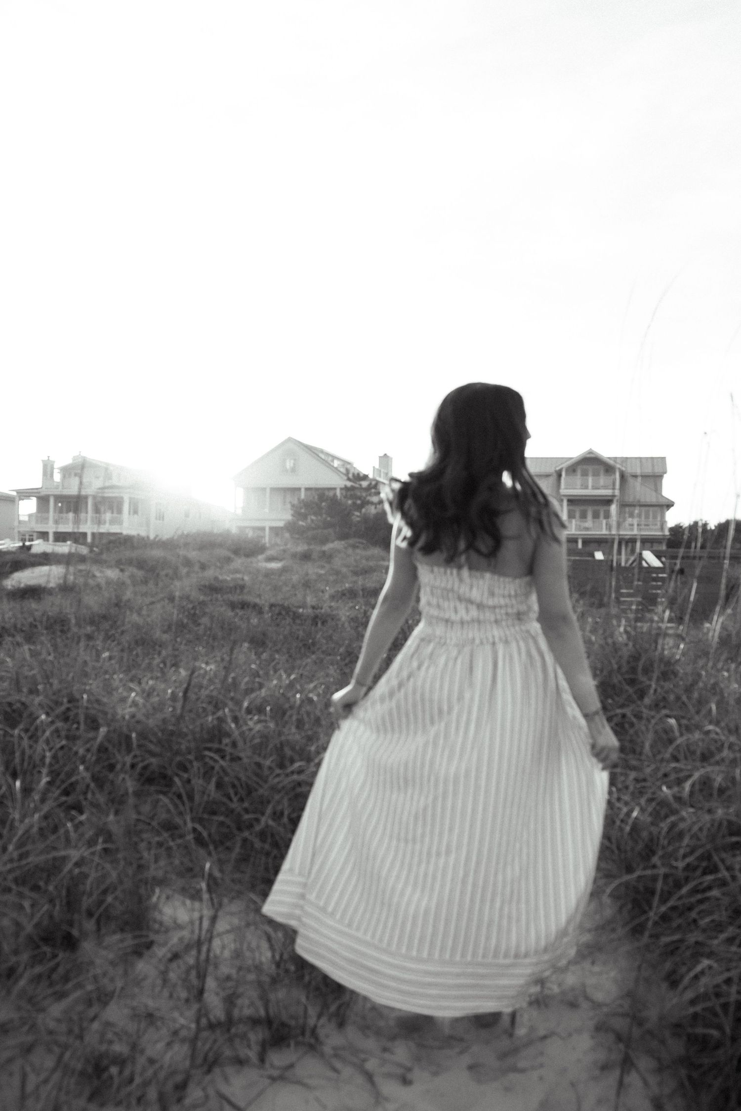 Black and white photo of a woman in a long striped dress walking toward beach houses on a sandy dune path.