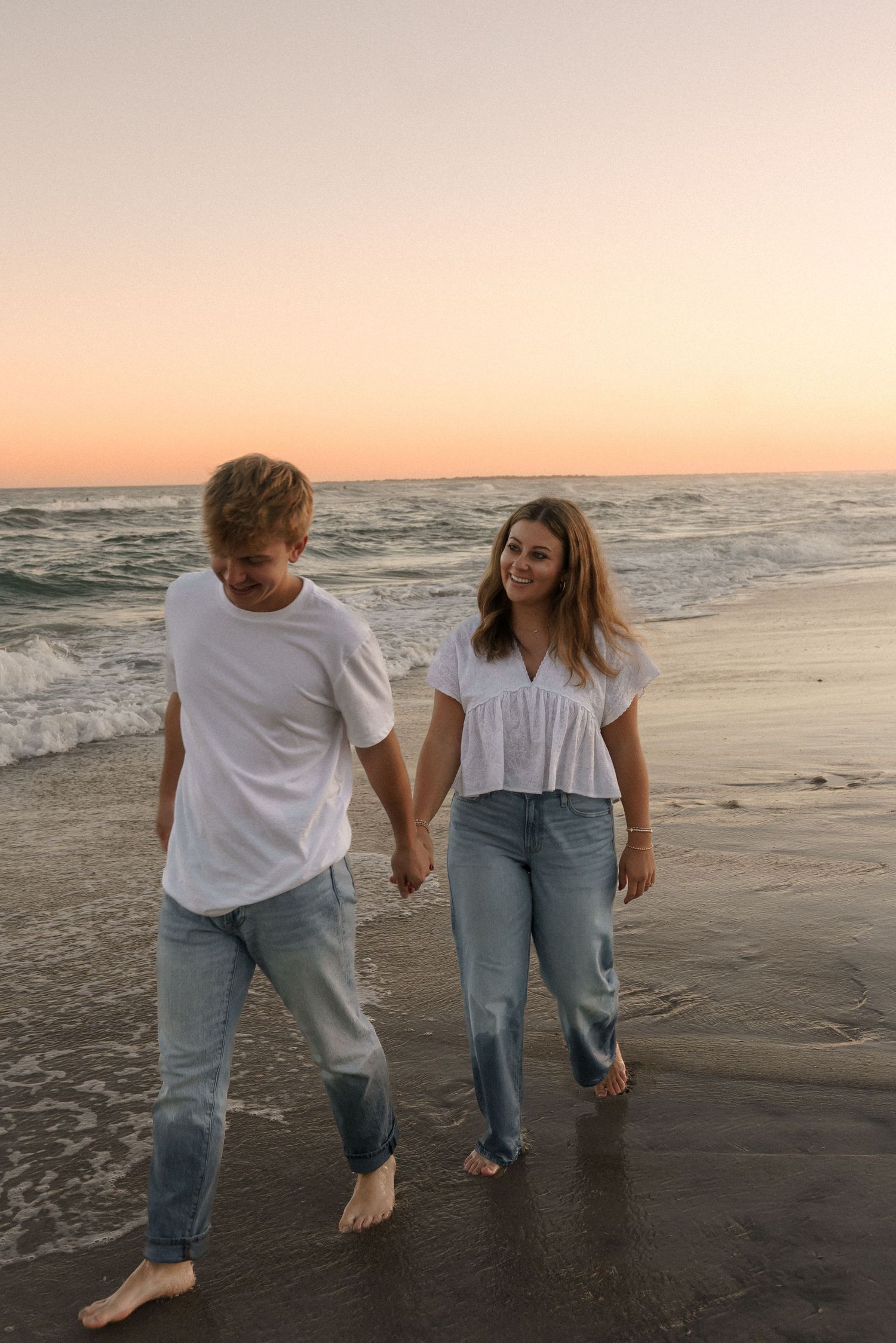 A couple walking hand in hand along the shoreline at sunset.