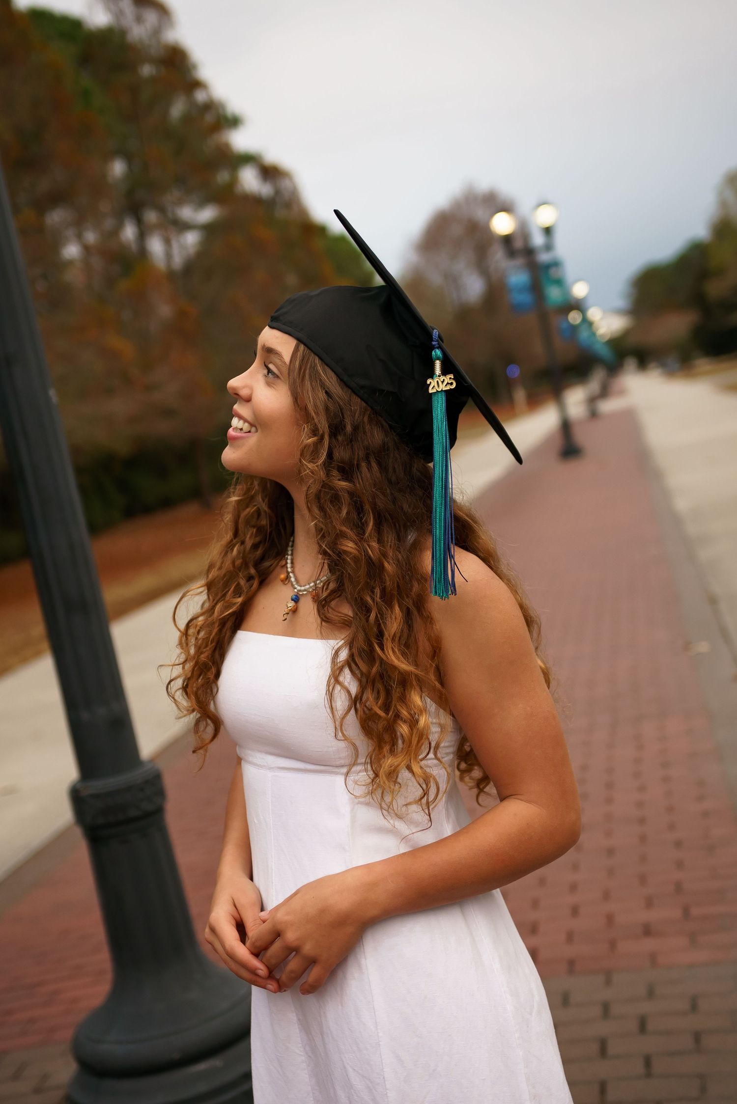 Graduate in a white dress and cap poses along a brick pathway during sunset.