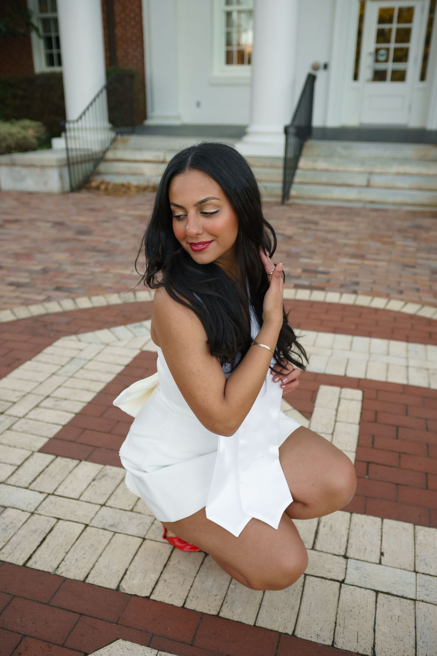 Woman in white dress kneeling gracefully on brick courtyard outside elegant building.