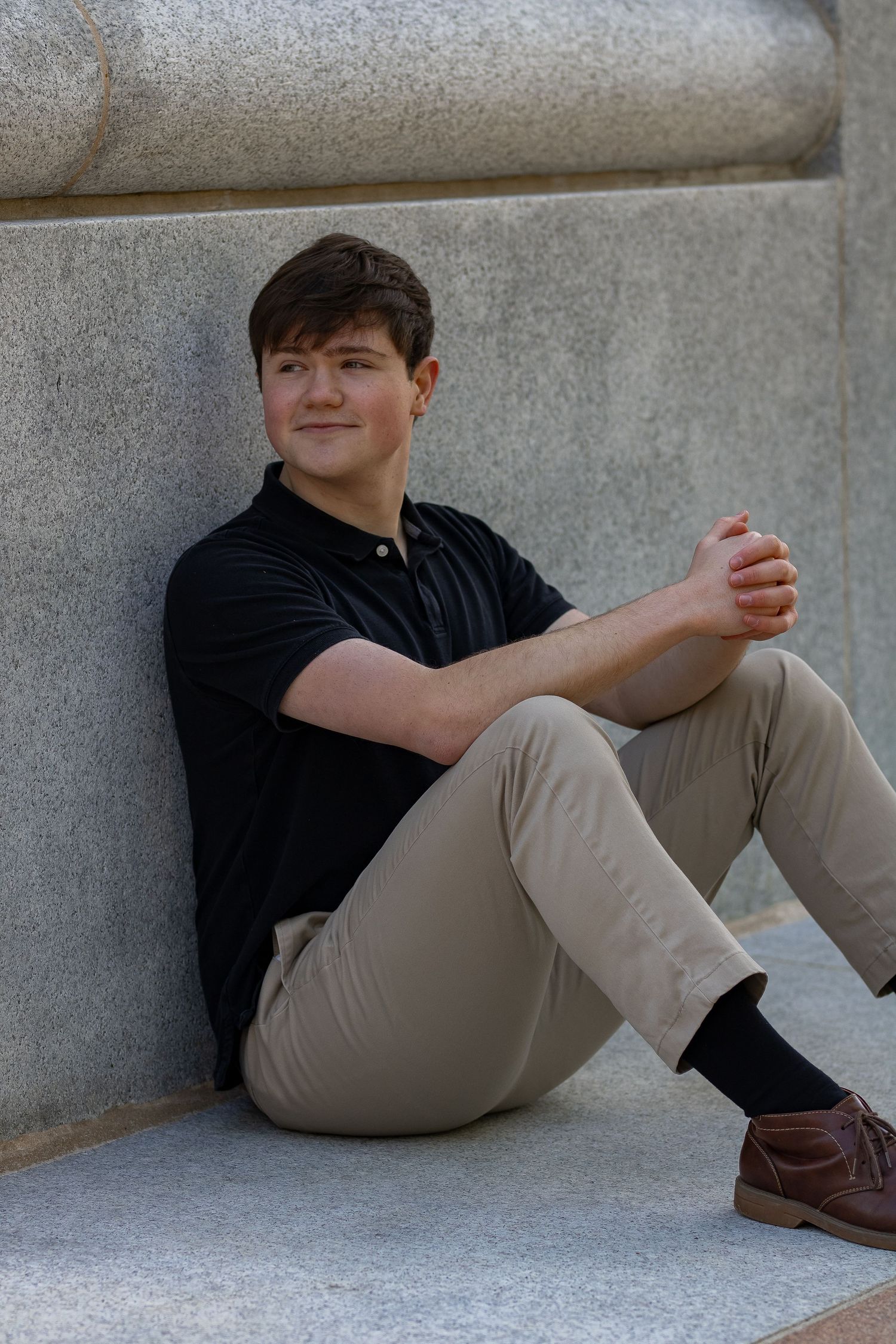 Student in casual attire reclines against concrete wall during photo session.