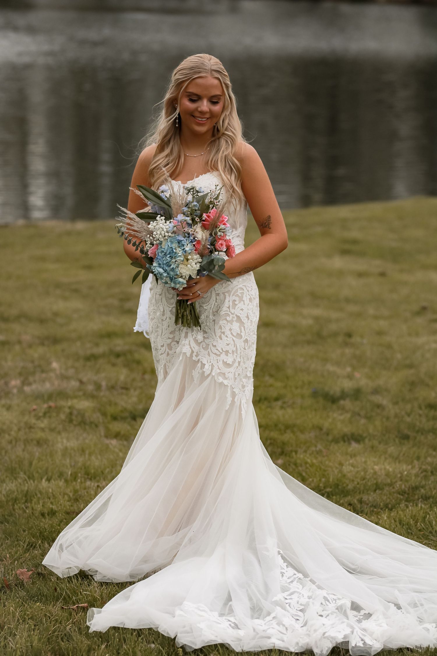 Bride in flowing white lace wedding dress holding colorful bouquet by lakeside.