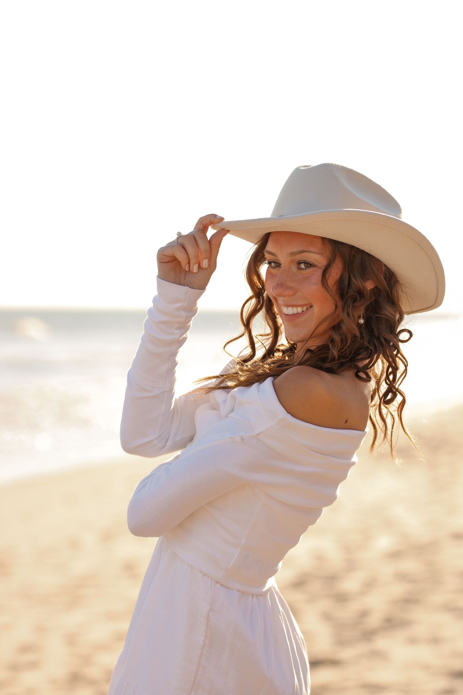 A person in a white dress and cowboy hat poses playfully on a sunny beach.