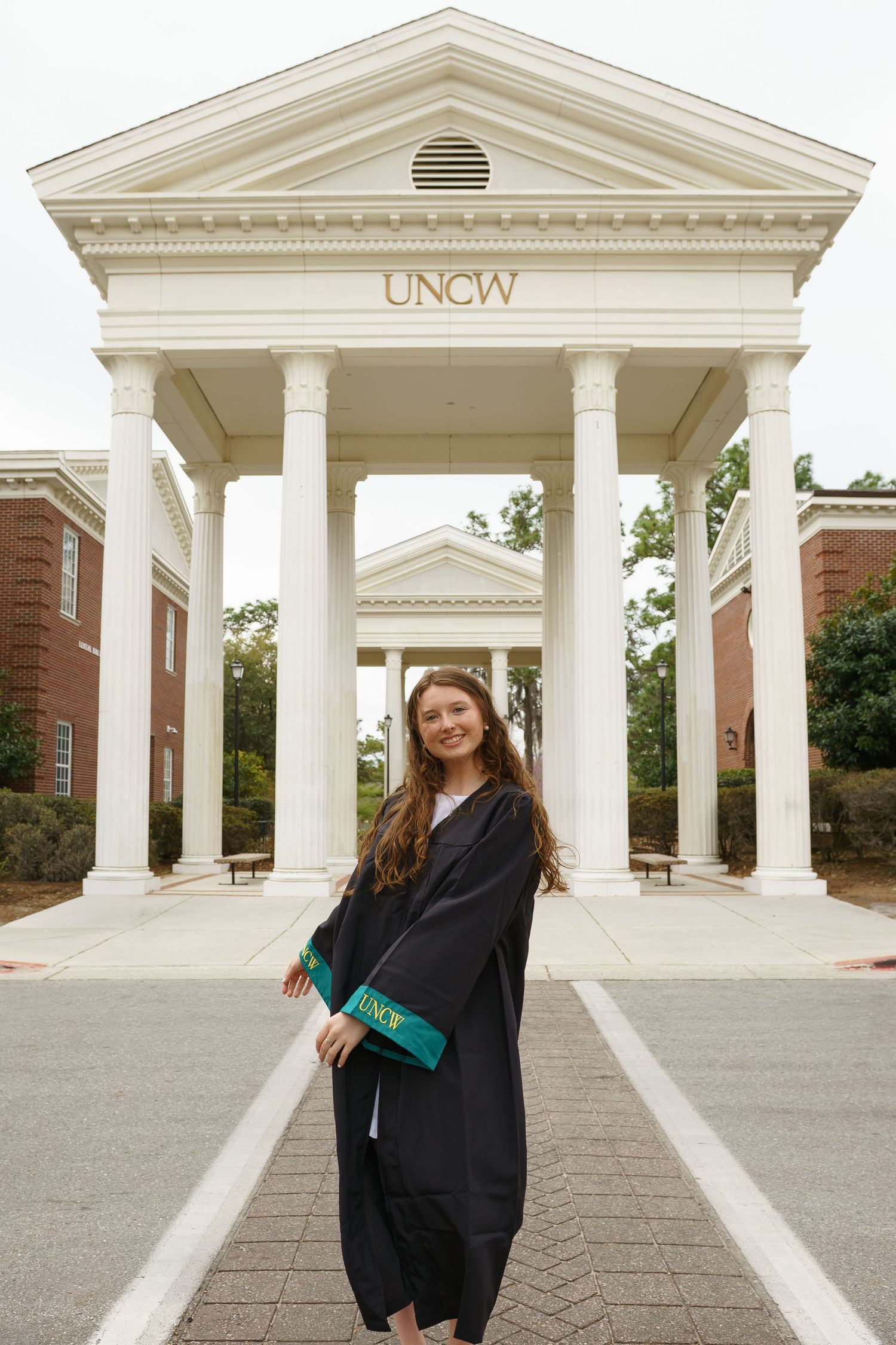 A smiling graduate in black cap and gown poses before the iconic UNCW columned entrance on campus.