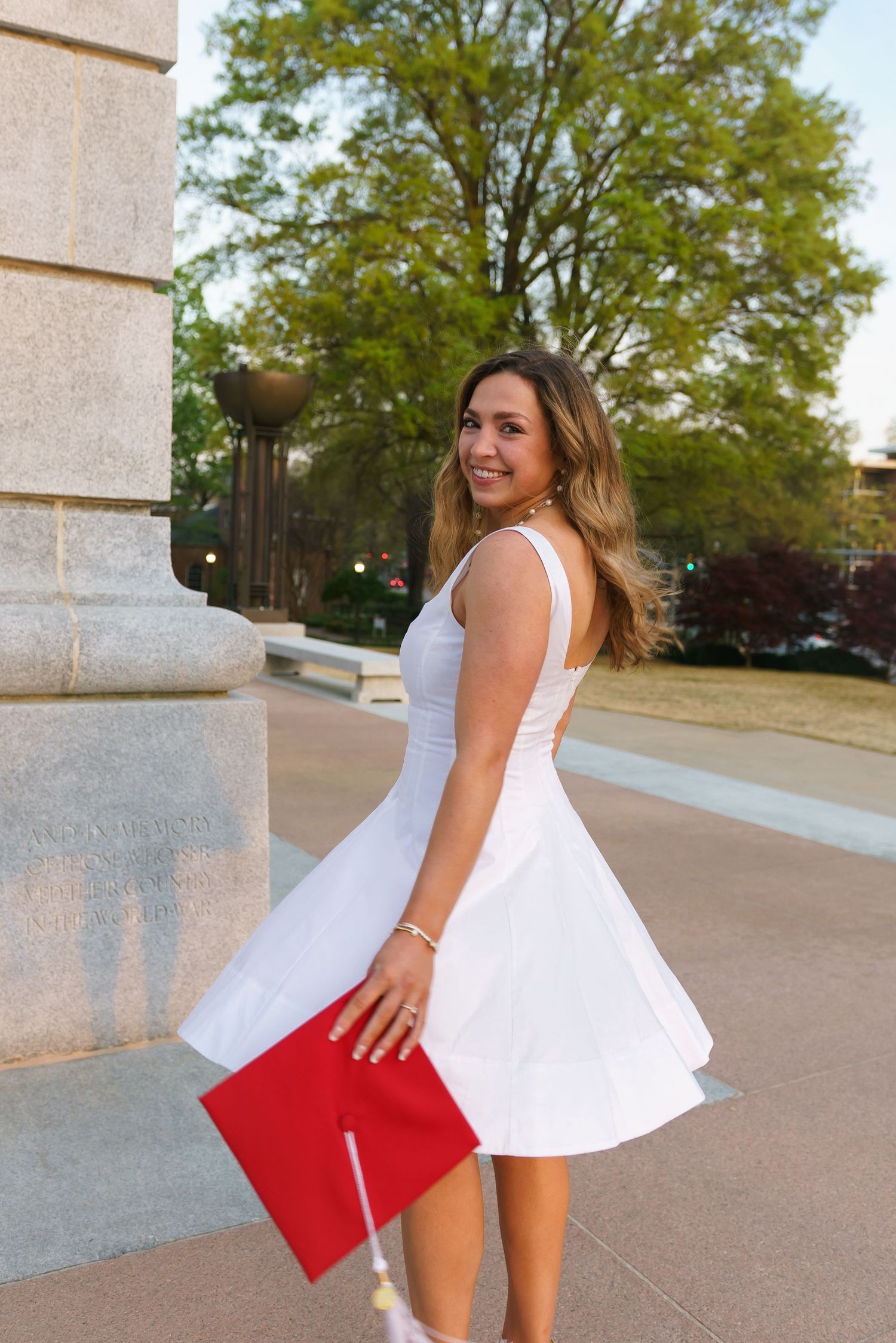 Young woman in white dress holding red graduation cap, smiling outdoors near stone column at university campus.