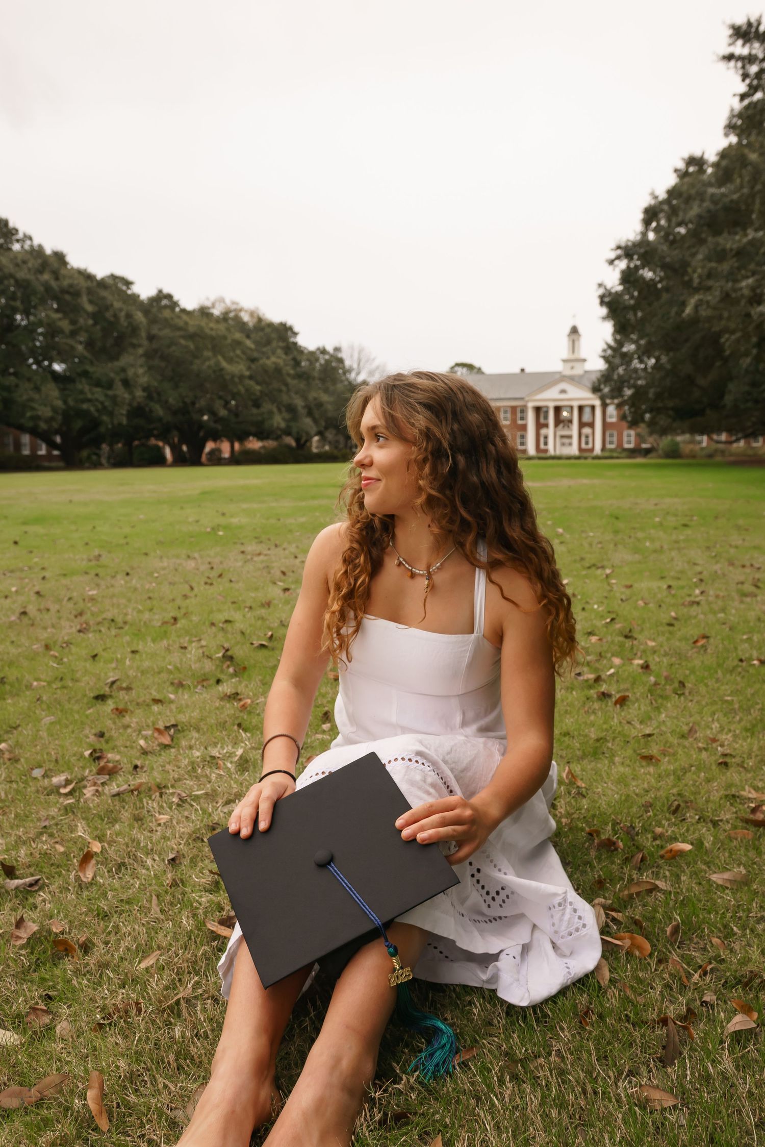 Graduate in white dress sits on lawn with UNCW buildings visible in background.