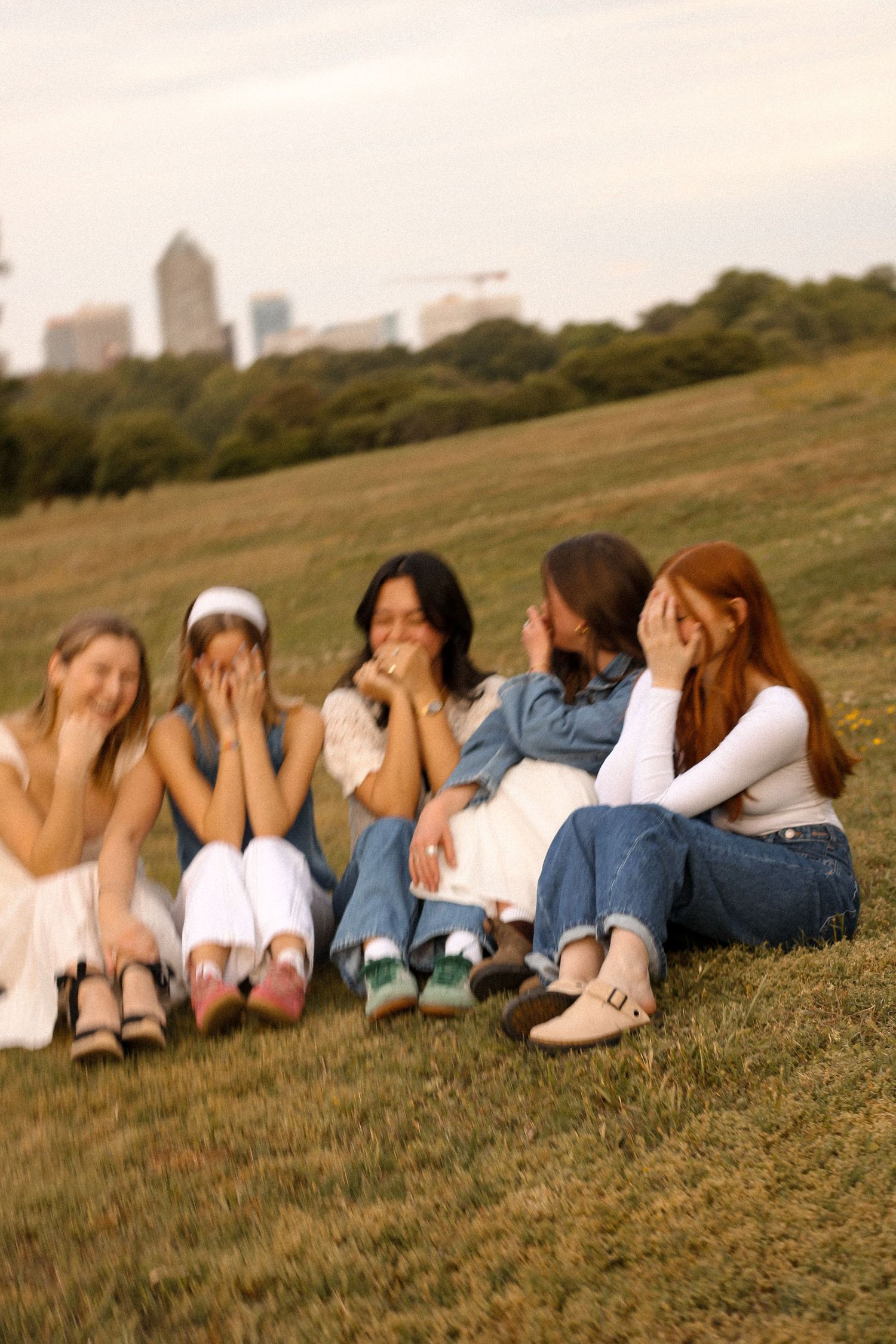 Friends lounging together on a grassy hill with city skyline visible in background at sunset.
